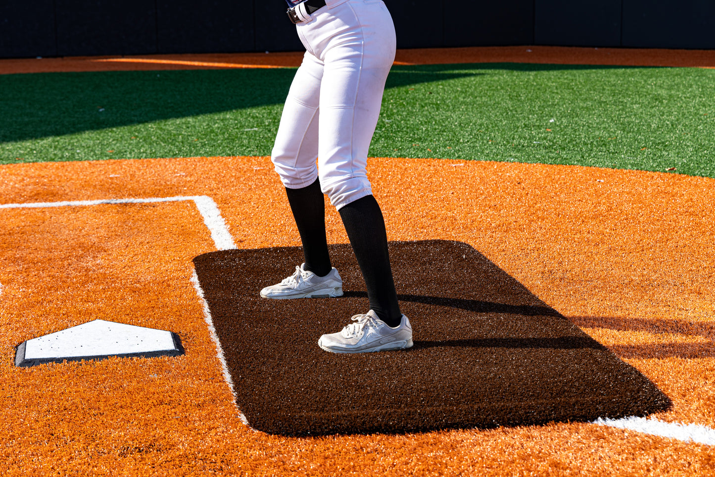A baseball player is positioned in the batter’s box near home plate on an orange and green artificial turf field, standing on the Portolite Half Batter’s Mat | 7′6″ x 3′6″ No Slip Batter’s Mat. Only the legs and lower torso are visible.