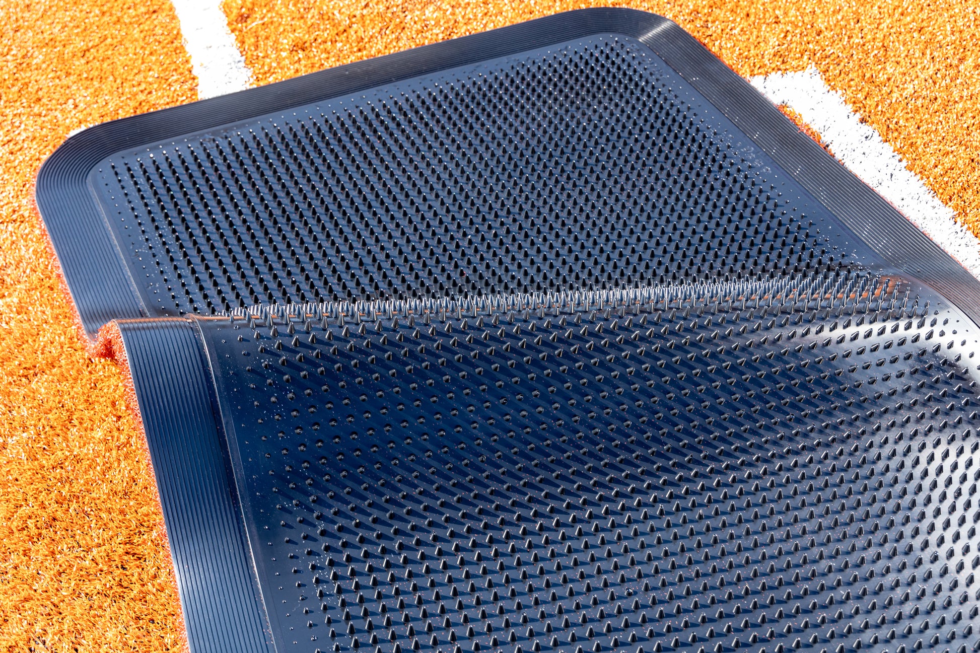 A close-up of the Portolite Half Batter’s Mat (7′6″ x 3′6″), a black no-slip rubber batting practice mat with small raised bumps, shown partially folded over on an orange textured surface with white lines—possibly a sports track or field. This portable mat is designed for durability and optimal performance during training sessions.