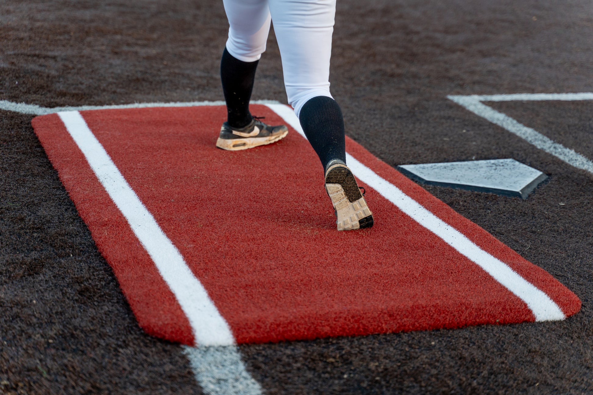 A baseball player in white pants and black cleats stands on a Portolite Half Batter’s Mat | 7′6″ x 3′6″ No Slip Batter’s Mat, featuring SpikeTek technology and white lines, placed on the brown dirt field near home plate.
