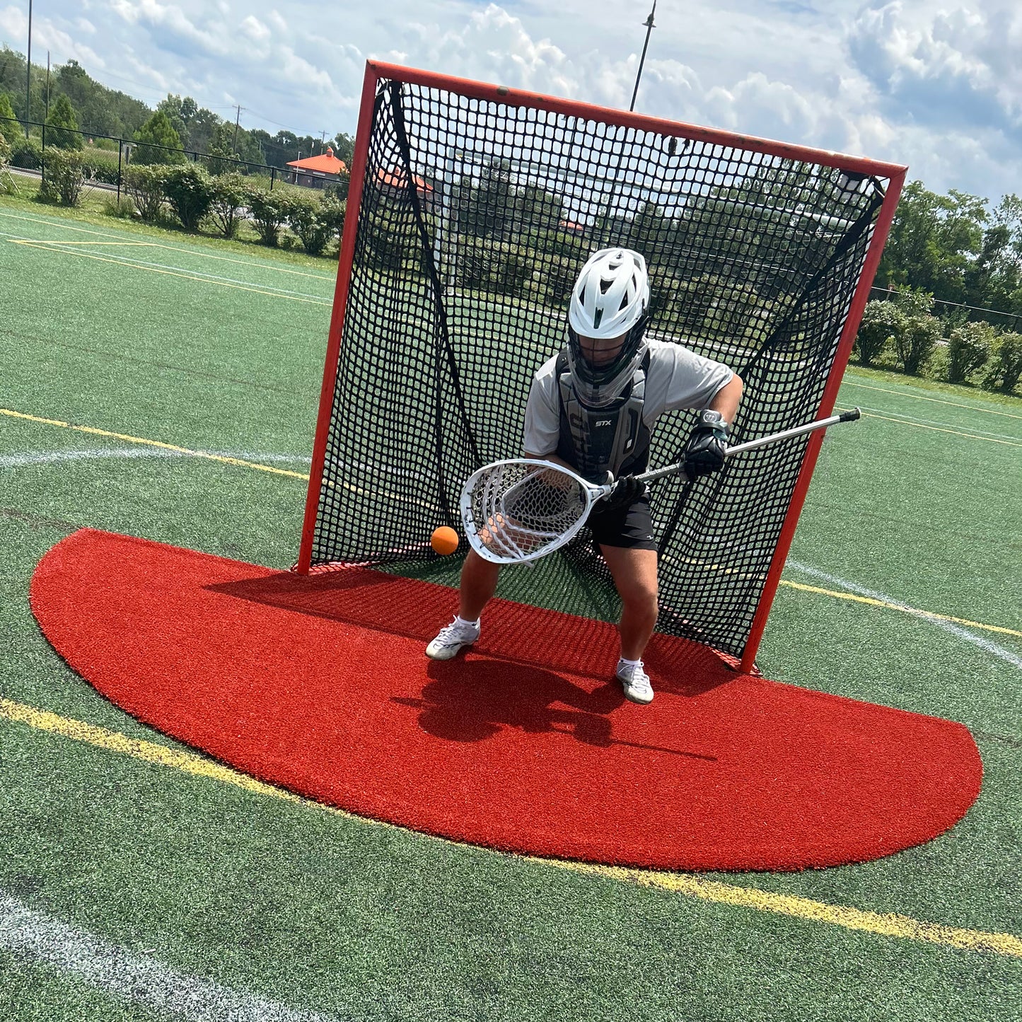 A lacrosse goalie in full gear crouches in front of a net, holding a stick to block an orange ball. The scene is on an outdoor turf field under a partly cloudy sky, where the Portolite Lacrosse Mat | Spiketek Turf Protection Mat For Practice and Drills provides extra turf protection during intense lacrosse practice.