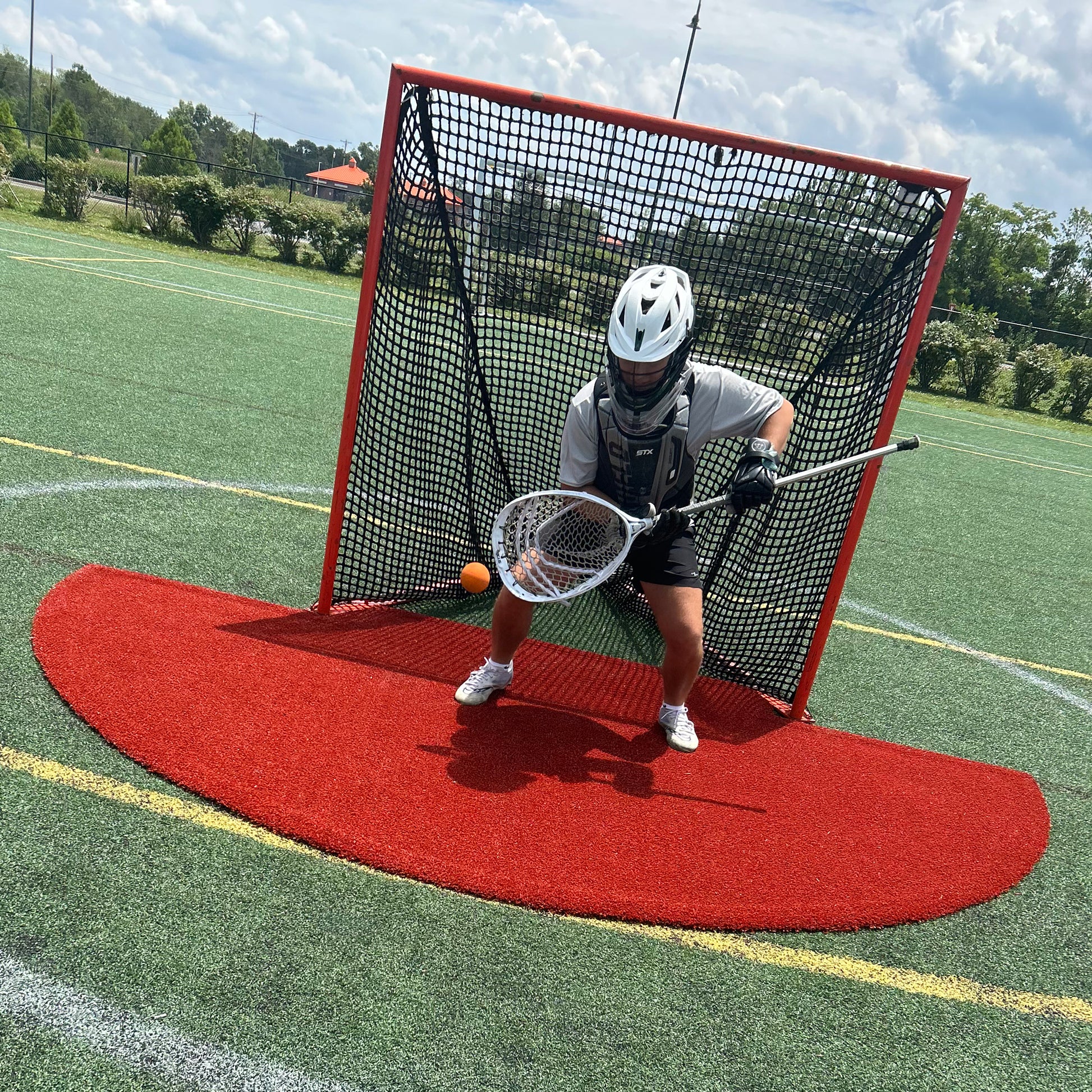 A lacrosse goalie in full gear crouches in front of a net, holding a stick to block an orange ball. The scene is on an outdoor turf field under a partly cloudy sky, where the Portolite Lacrosse Mat | Spiketek Turf Protection Mat For Practice and Drills provides extra turf protection during intense lacrosse practice.