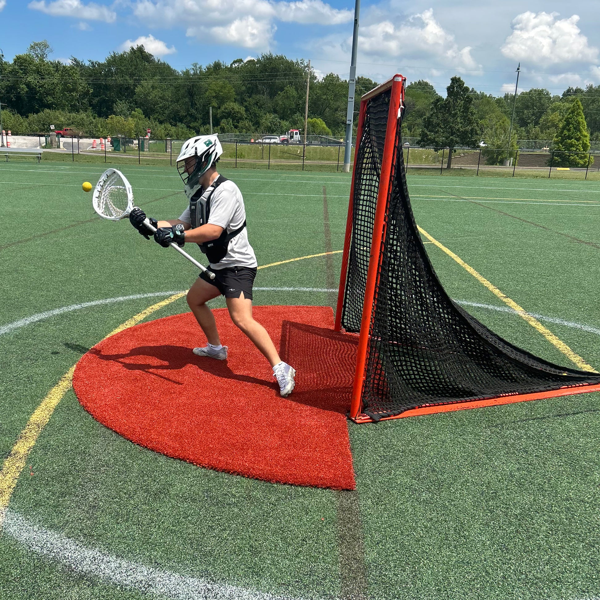 A lacrosse goalie in gear catches a ball with their stick in front of the net on a green outdoor field under a partly cloudy sky, demonstrating essential skills during practice while benefiting from turf protection provided by the Portolite Lacrosse Mat | Spiketek Turf Protection Mat for Practice and Drills.