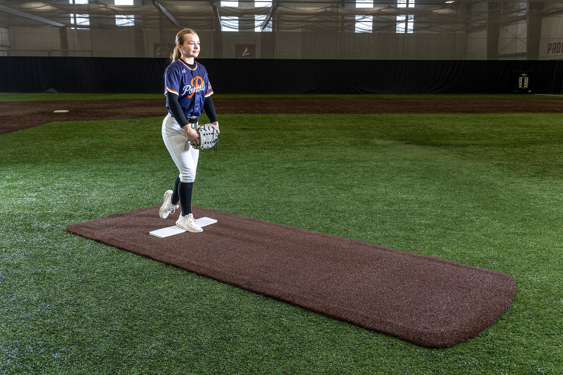 A softball player in uniform stands on the pitcher’s mound inside an indoor field, holding a glove and appearing ready to pitch. She stands on the Portolite Pro Spiked Game Mat No Lines / Lanes | Collegiate & Pro-Level Pitching, which provides turf protection beneath her feet. The facility is well-lit with artificial turf and netting in the background.
