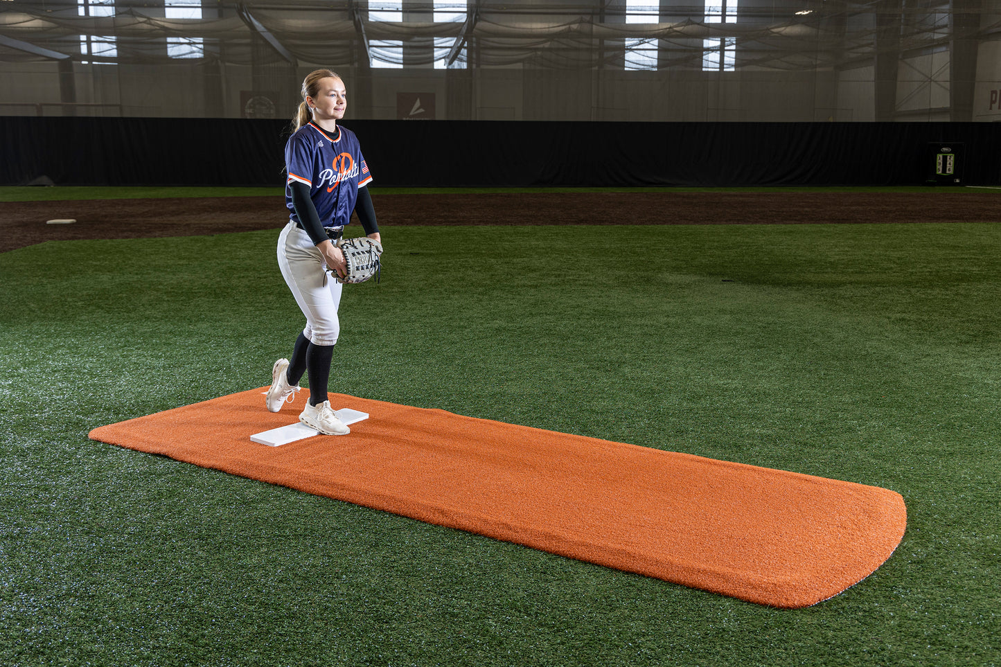 A softball player in uniform balances on one leg with a glove and ball, preparing to pitch indoors on the Portolite Pro Spiked Game Mat No Lines / Lanes, designed for collegiate and pro-level pitching.