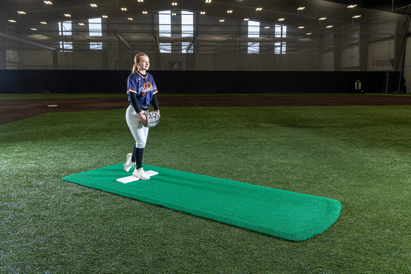 A young female softball pitcher stands on a Portolite Pro Spiked Game Mat No Lines / Lanes | Collegiate & Pro-Level Pitching indoors, wearing a blue jersey and holding a glove, inside a large turf training facility designed for optimal turf protection.