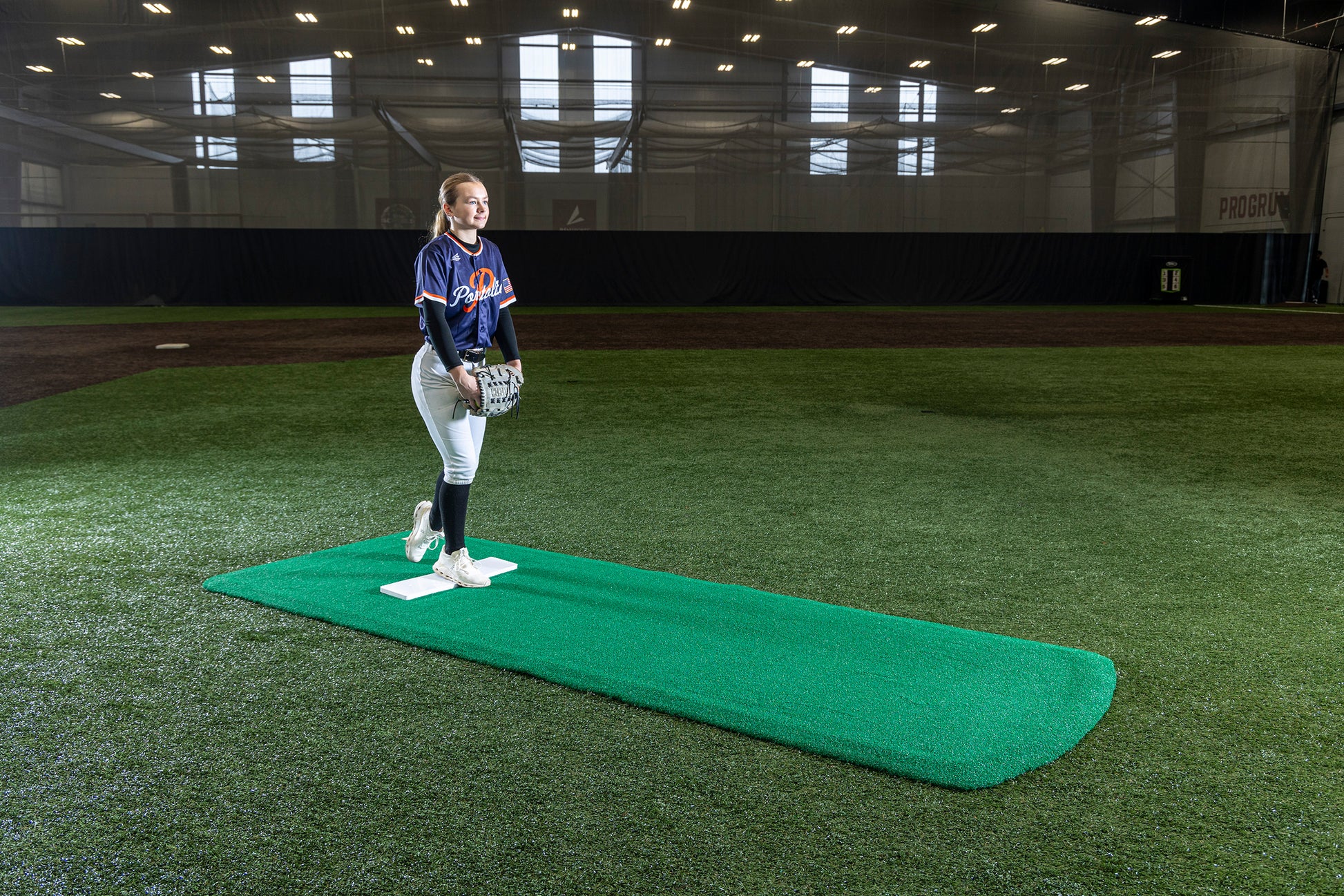 A young female softball pitcher stands on a Portolite Pro Spiked Game Mat No Lines / Lanes | Collegiate & Pro-Level Pitching indoors, wearing a blue jersey and holding a glove, inside a large turf training facility designed for optimal turf protection.