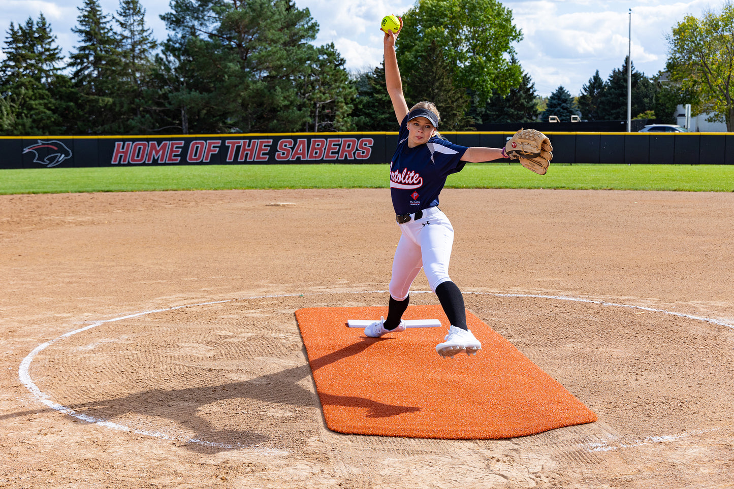 A softball pitcher in a navy jersey and white pants winds up to throw a pitch on an orange mound, stepping confidently onto the Portolite Pro Spiked Game Mat No Lines / Lanes | Collegiate & Pro-Level Pitching for added turf protection, with a HOME OF THE SABERS banner in the background.