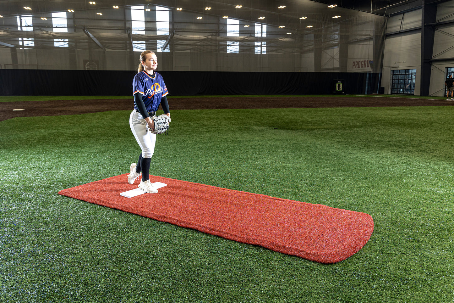 A softball player stands on a Portolite Pro Spiked Game Mat No Lines / Lanes | Collegiate & Pro-Level Pitching inside an indoor field, holding a ball and glove, ready to pitch. Sunlight streams through large windows behind her, highlighting the turf protection provided by the long spiked game mat beneath her feet.