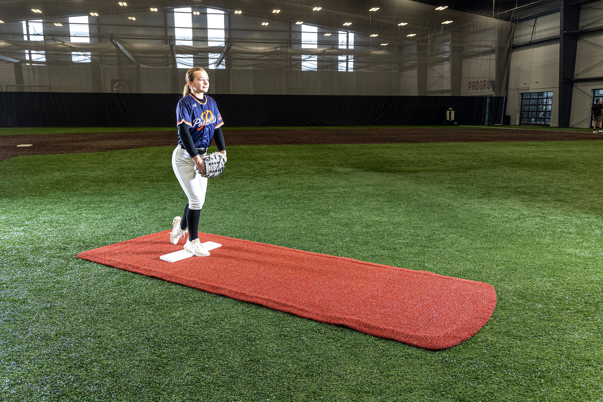 A softball player stands on a Portolite Pro Spiked Game Mat No Lines / Lanes | Collegiate & Pro-Level Pitching inside an indoor field, holding a ball and glove, ready to pitch. Sunlight streams through large windows behind her, highlighting the turf protection provided by the long spiked game mat beneath her feet.