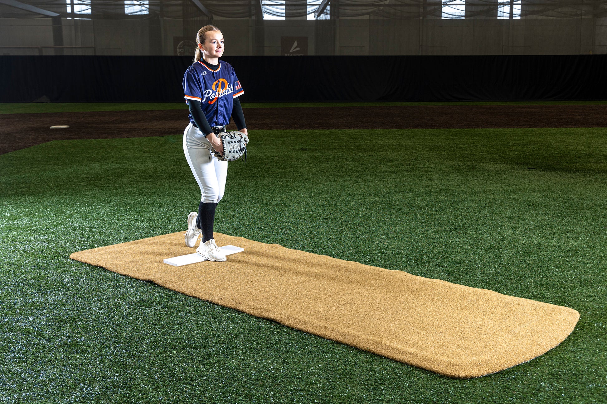 A softball player in uniform stands on a pitching mound indoors, holding a glove and softball and preparing to pitch. The Portolite Pro Spiked Game Mat No Lines / Lanes | Collegiate & Pro-Level Pitching provides turf protection underfoot, while artificial turf and dark netting are visible in the background.