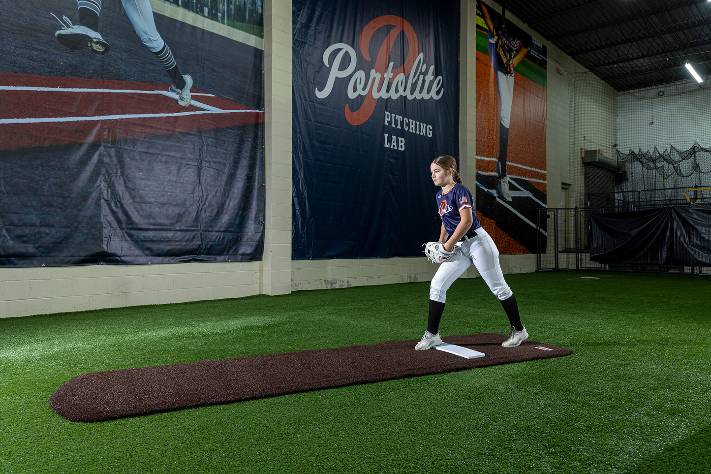 A softball player in uniform stands on a Portolite Signature Practice Mat with Spikes No Lines / Lanes | 12U Pitchers, positioned on an indoor pitching mound at a training facility. Large banners and synthetic turf are visible in the background, creating the ideal environment to practice softball pitching.