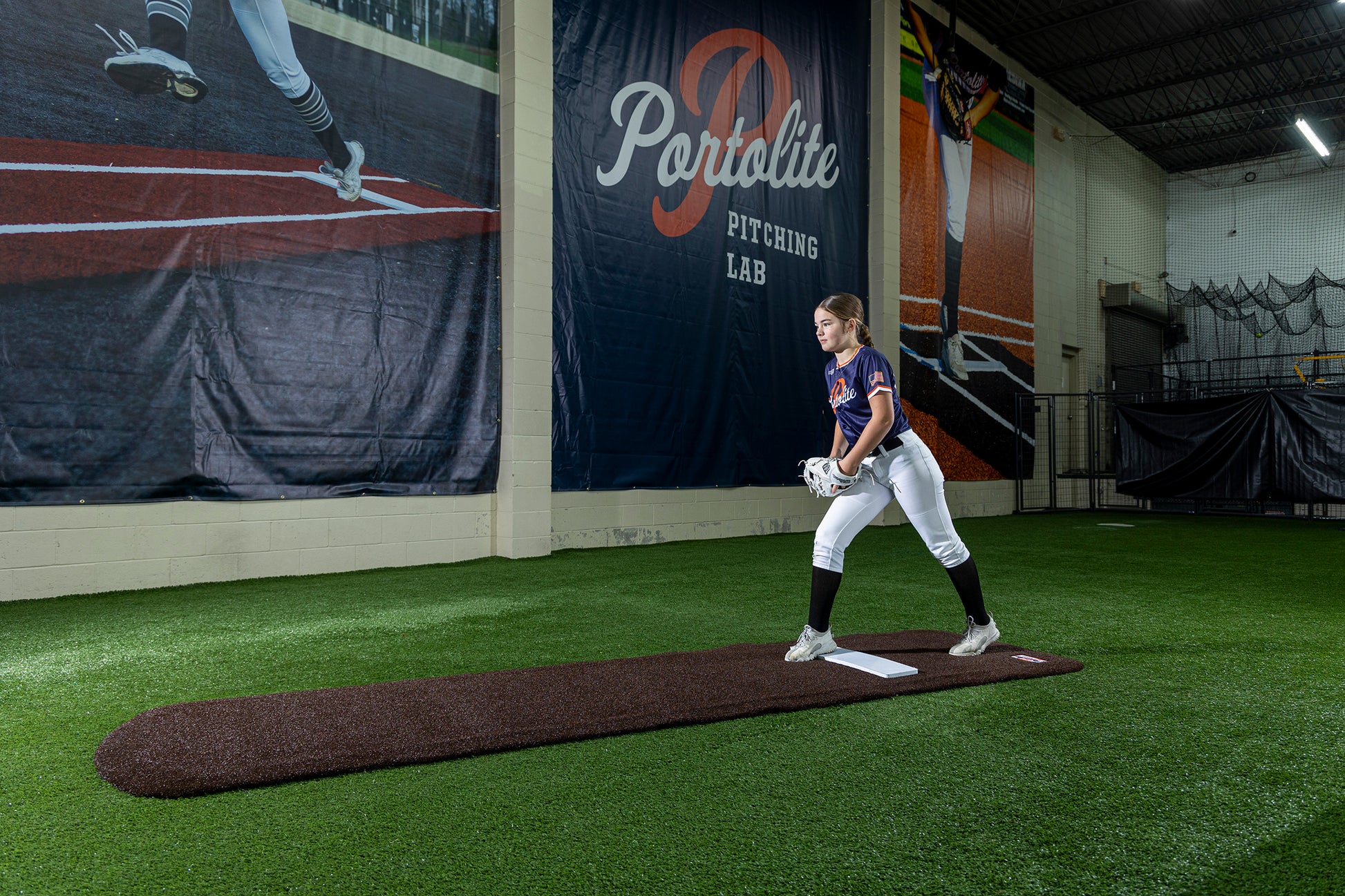A softball player in uniform stands on a Portolite Signature Practice Mat with Spikes No Lines / Lanes | 12U Pitchers, positioned on an indoor pitching mound at a training facility. Large banners and synthetic turf are visible in the background, creating the ideal environment to practice softball pitching.