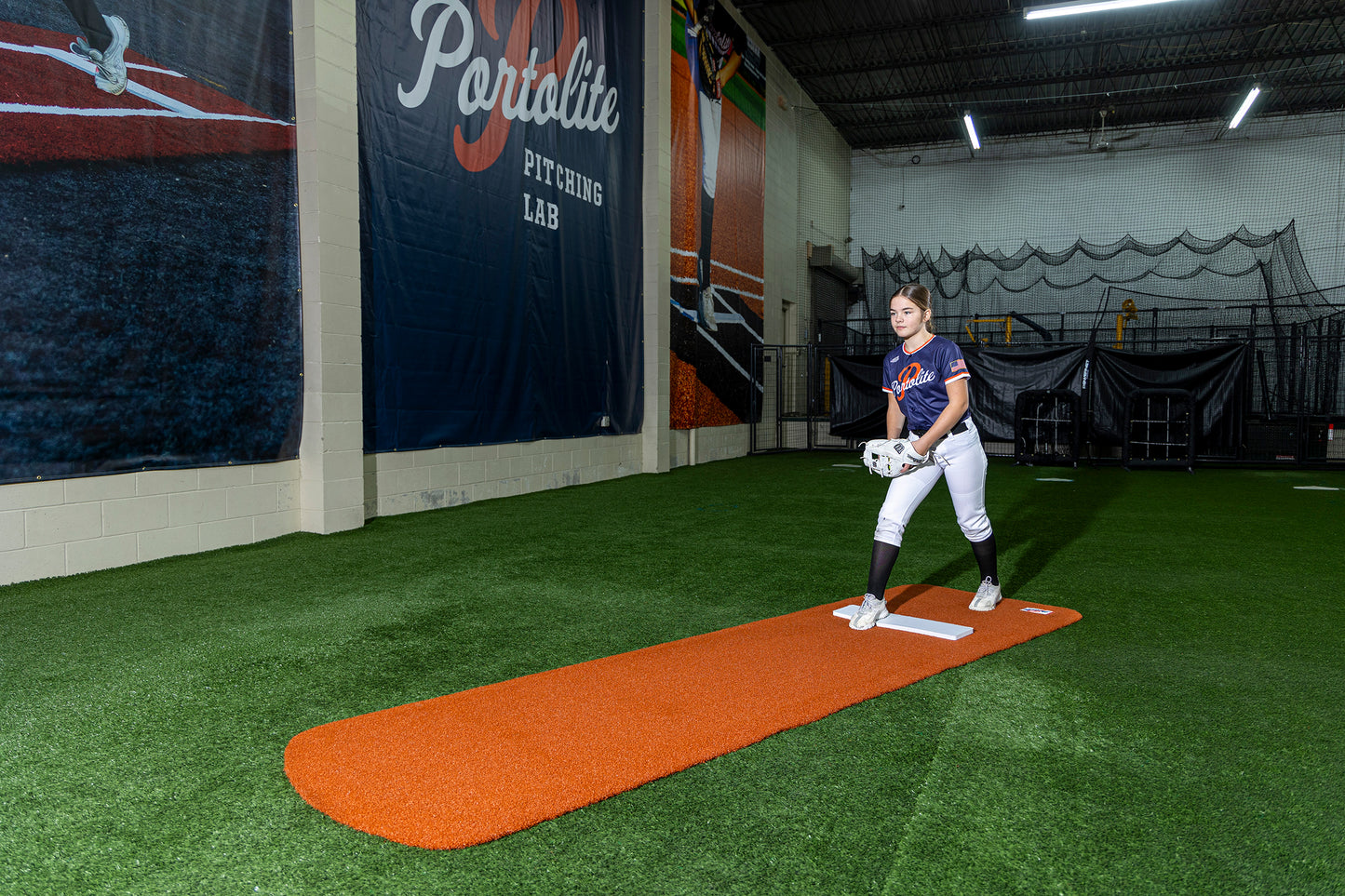 A softball player in uniform stands on a Portolite Signature Practice Mat with Spikes No Lines / Lanes | 12U Pitchers inside an indoor training facility with synthetic turf flooring and large banners on the walls, ready to practice softball pitching.