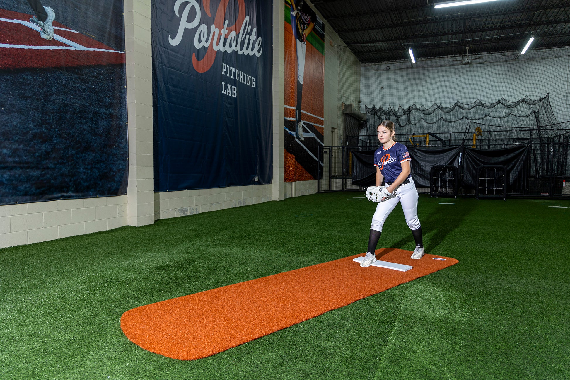 A softball player in uniform stands on a Portolite Signature Practice Mat with Spikes No Lines / Lanes | 12U Pitchers inside an indoor training facility with synthetic turf flooring and large banners on the walls, ready to practice softball pitching.