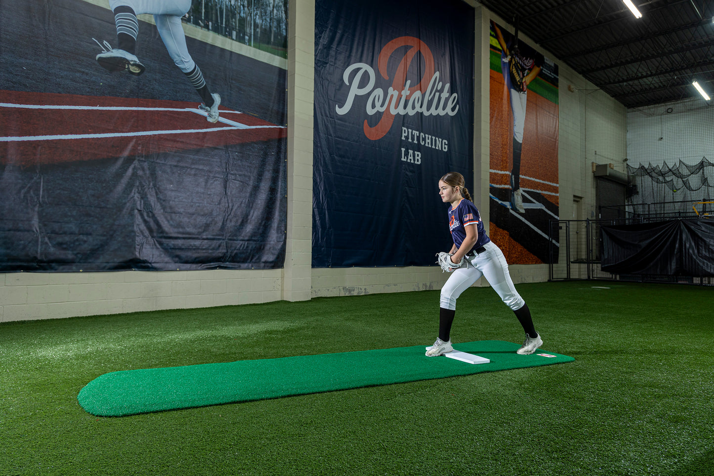 A young softball player stands indoors on the Portolite Signature Practice Mat with Spikes No Lines / Lanes | 12U Pitchers, preparing to pitch on the green synthetic turf. Large banners featuring baseball imagery and the words “Portolite Pitching Lab” hang on the wall behind her, creating the perfect atmosphere to practice softball pitching.