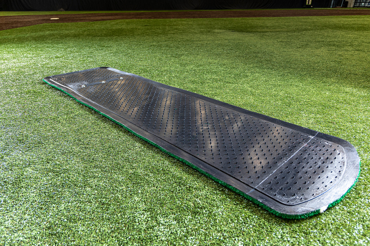 A Portolite Signature Practice Mat with Spikes No Lines / Lanes | 12U Pitchers, featuring a black rubber textured surface for softball pitching practice, sits on green synthetic turf in an indoor facility.