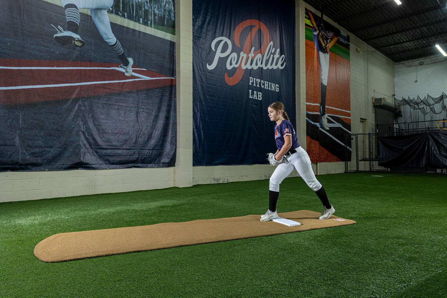 A young softball player stands on a pitching mound in an indoor training facility, preparing to practice with the Portolite Signature Practice Mat with Spikes No Lines / Lanes | 12U Pitchers. Large banners hang overhead, and synthetic turf covers the area, providing the perfect surface for training.
