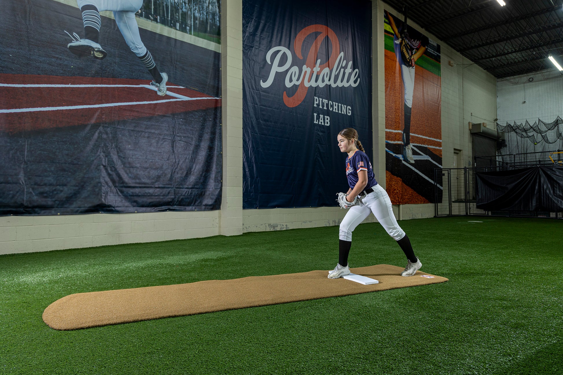 A young softball player stands on a pitching mound in an indoor training facility, preparing to practice with the Portolite Signature Practice Mat with Spikes No Lines / Lanes | 12U Pitchers. Large banners hang overhead, and synthetic turf covers the area, providing the perfect surface for training.