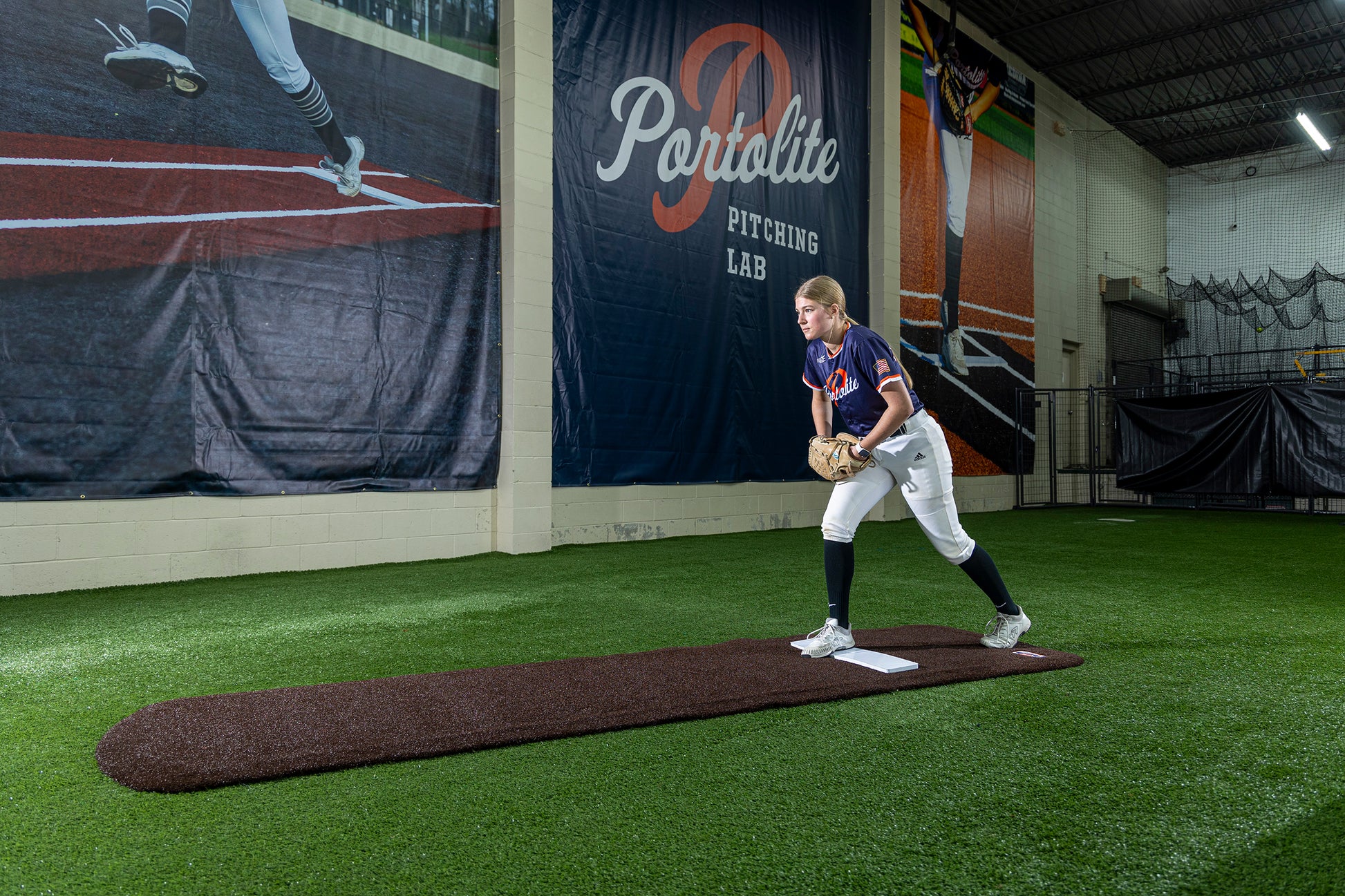 A softball player stands on an indoor pitching mound, preparing to pitch. Large banners on the wall read Portolite Pitching Lab. The facility features artificial turf, the Portolite Ultimate Spiked Practice Mat No Lanes / Lines for bullpen practice—suitable for all ages and skill levels—and sports equipment throughout.