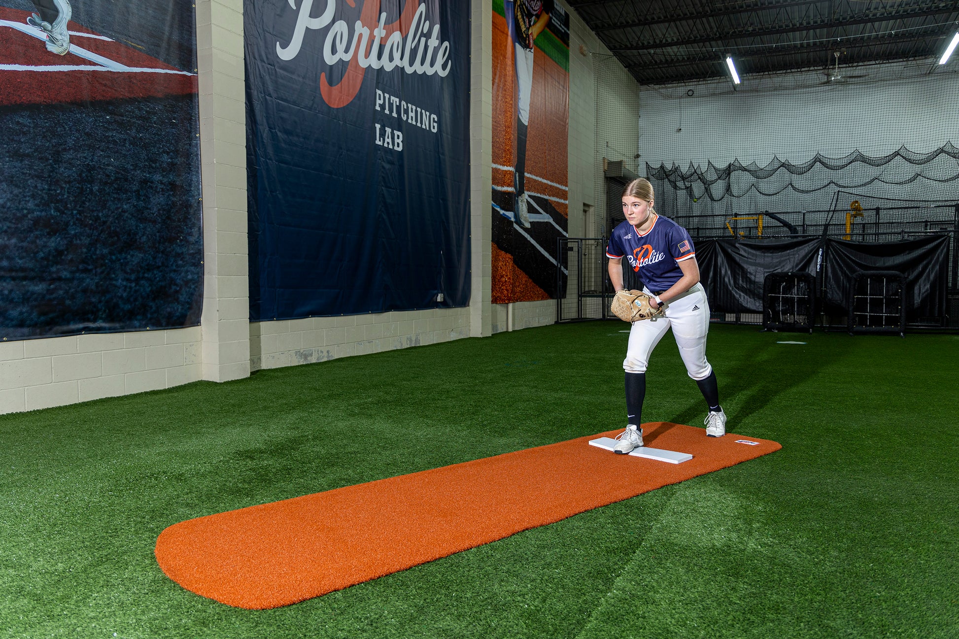 An indoor softball pitcher stands on the orange mound, wearing a blue and red uniform with white pants, ready to throw. Behind her hangs a Portolite Pitching Lab banner over turf flooring. For her bullpen session, the Portolite Ultimate Spiked Practice Mat No Lanes / Lines (suitable for all ages and skill levels) is set up beneath her feet.