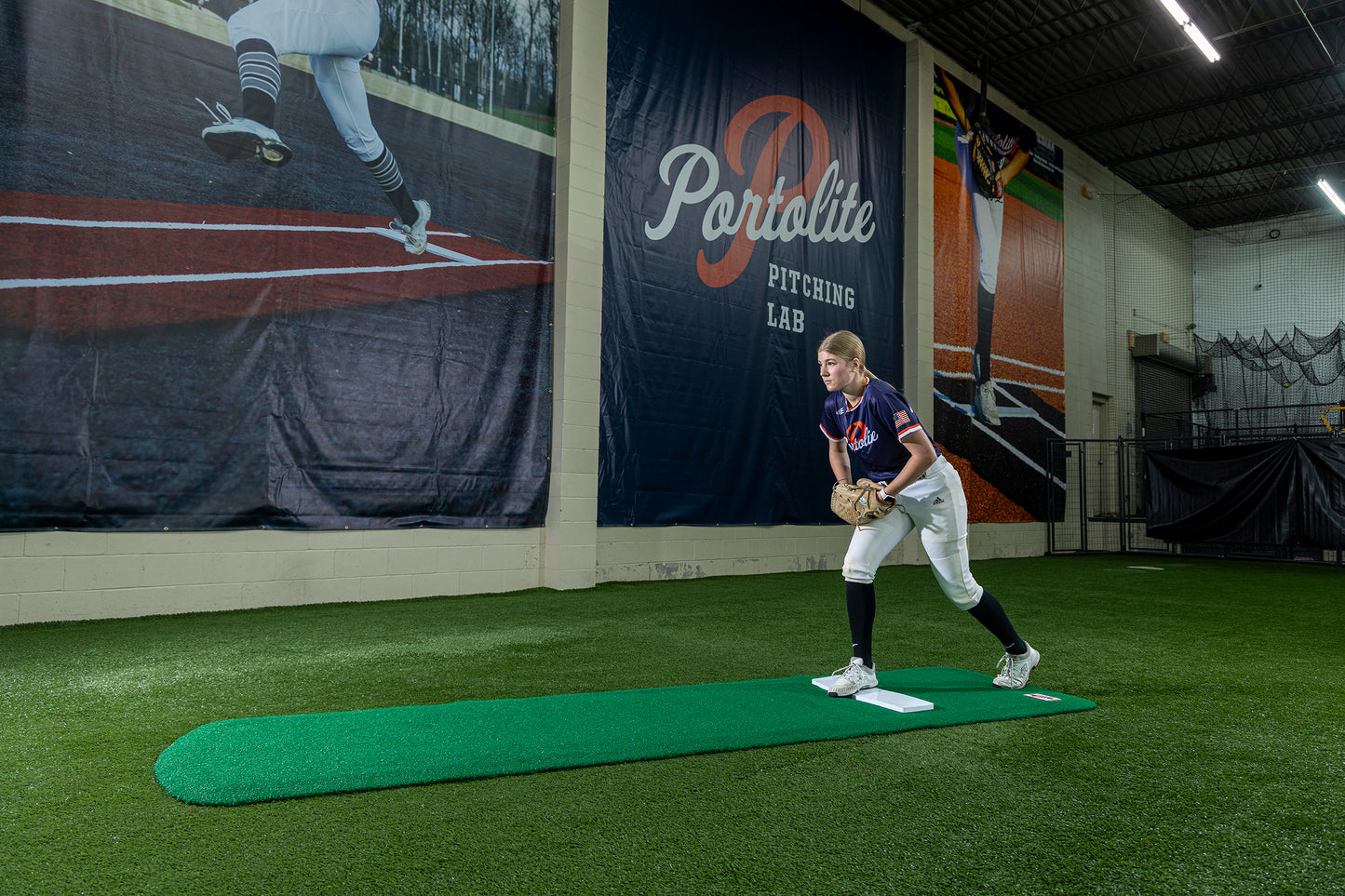 A baseball player stands on an artificial mound in an indoor pitching lab, holding a glove and preparing to pitch. Large baseball-themed wall graphics are visible behind him, and the Portolite Ultimate Spiked Practice Mat No Lanes / Lines | All Ages and Skill Levels enhances the bullpen practice environment.