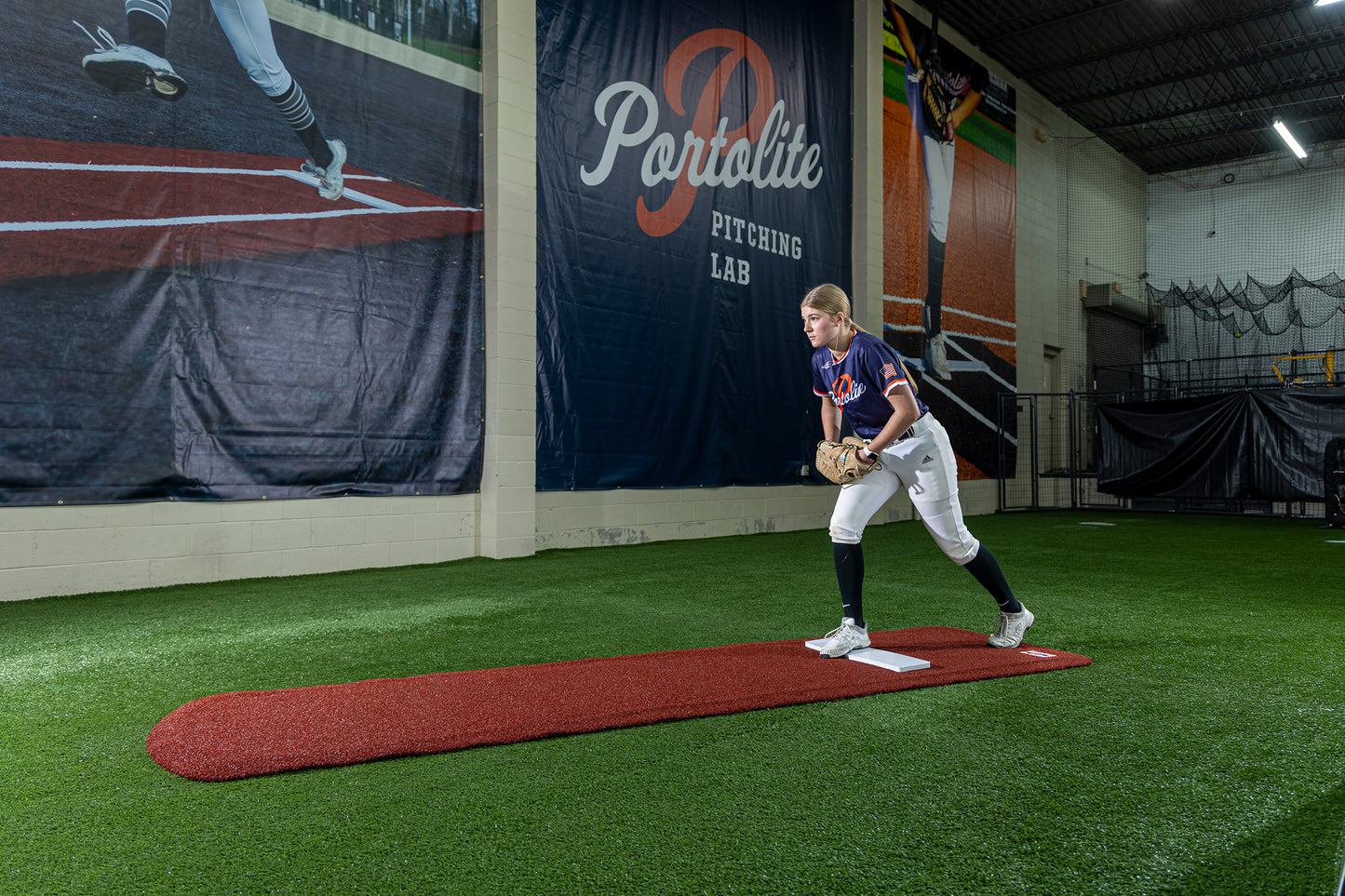 A softball player stands on a red pitching mound in an indoor training facility, preparing to pitch. The wall behind displays a large Portolite Pitching Lab sign, while the Portolite Ultimate Spiked Practice Mat No Lanes / Lines | All Ages and Skill Levels enhances the surface for bullpen practice.