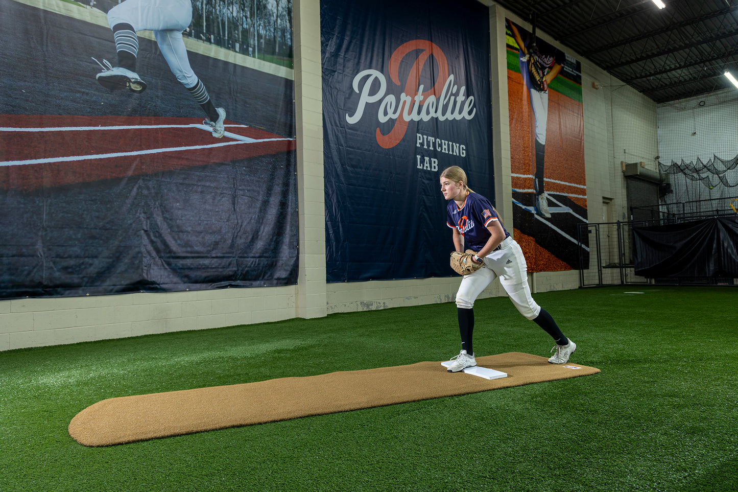 A baseball player in uniform practices pitching on a mound indoors at Portolite Pitching Lab, with a large mural showing a pitcher in the background. The Portolite Ultimate Spiked Practice Mat No Lanes / Lines, designed for all ages and skill levels, provides enhanced stability during bullpen sessions, ensuring each pitch is delivered with precision.