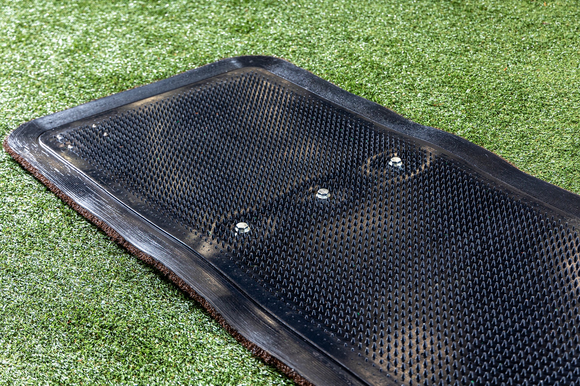 Close-up of the Portolite Ultimate Spiked Practice Mat No Lanes / Lines, featuring a black textured rubber surface secured with three bolts, resting on artificial green grass. The rectangular mat is designed for all ages and skill levels, making it ideal for bullpen practice or outdoor sports use.