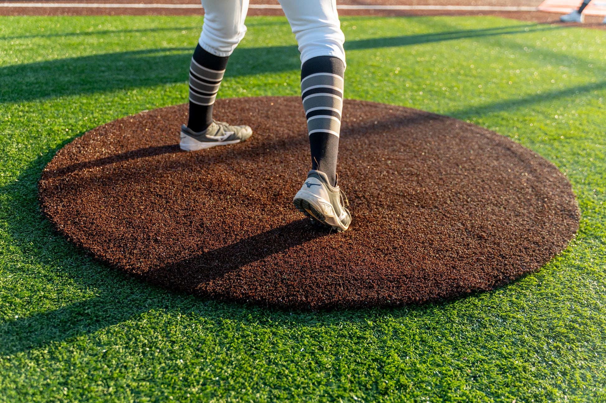 A baseball player stands on a pitching mound, wearing cleats, striped socks, and white pants, beside the Portolite On-Deck Circle on a synthetic turf baseball field with vibrant green artificial grass.