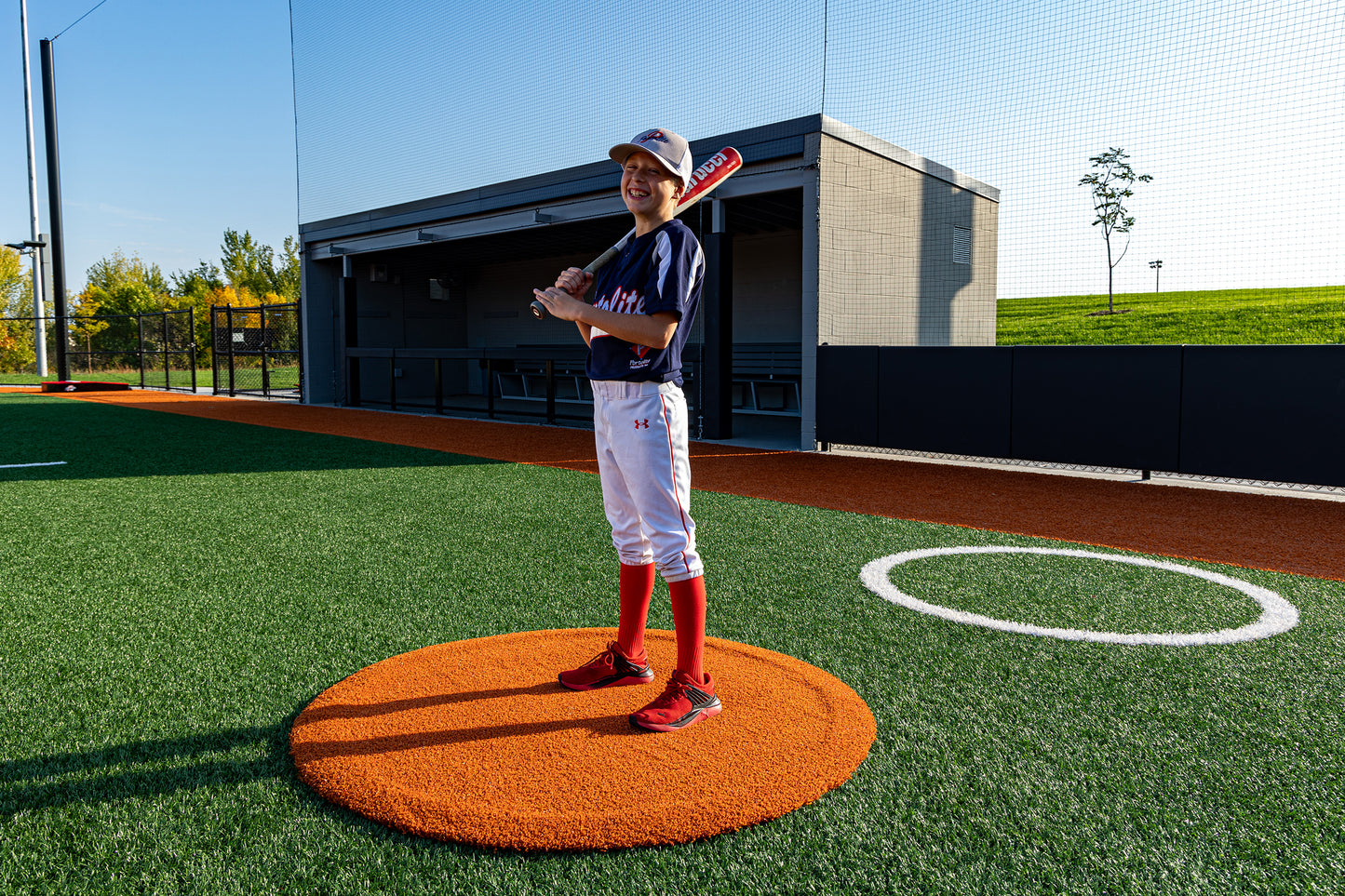 A young baseball player stands on a sunny, vibrant synthetic turf field, smiling and holding a bat. He wears a navy and white uniform with red accents while standing next to the Portolite On-Deck Circle, available in 4′, 5′, and 6′ sizes.
