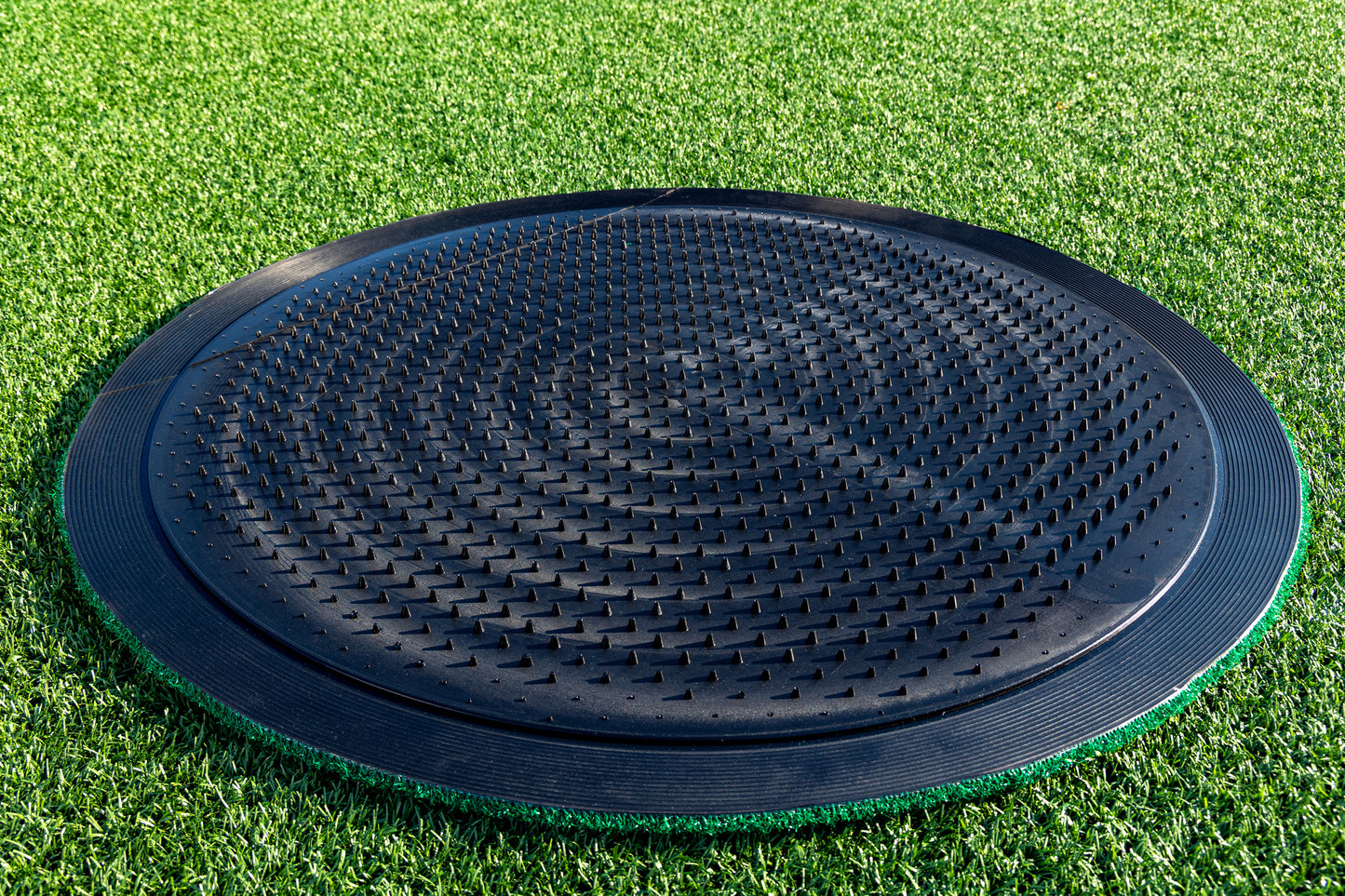 A Portolite On-Deck Circle featuring a large, black, textured plastic design with raised bumps is positioned on synthetic turf baseball grass, likely intended for balance or agility training near the on-deck area.
