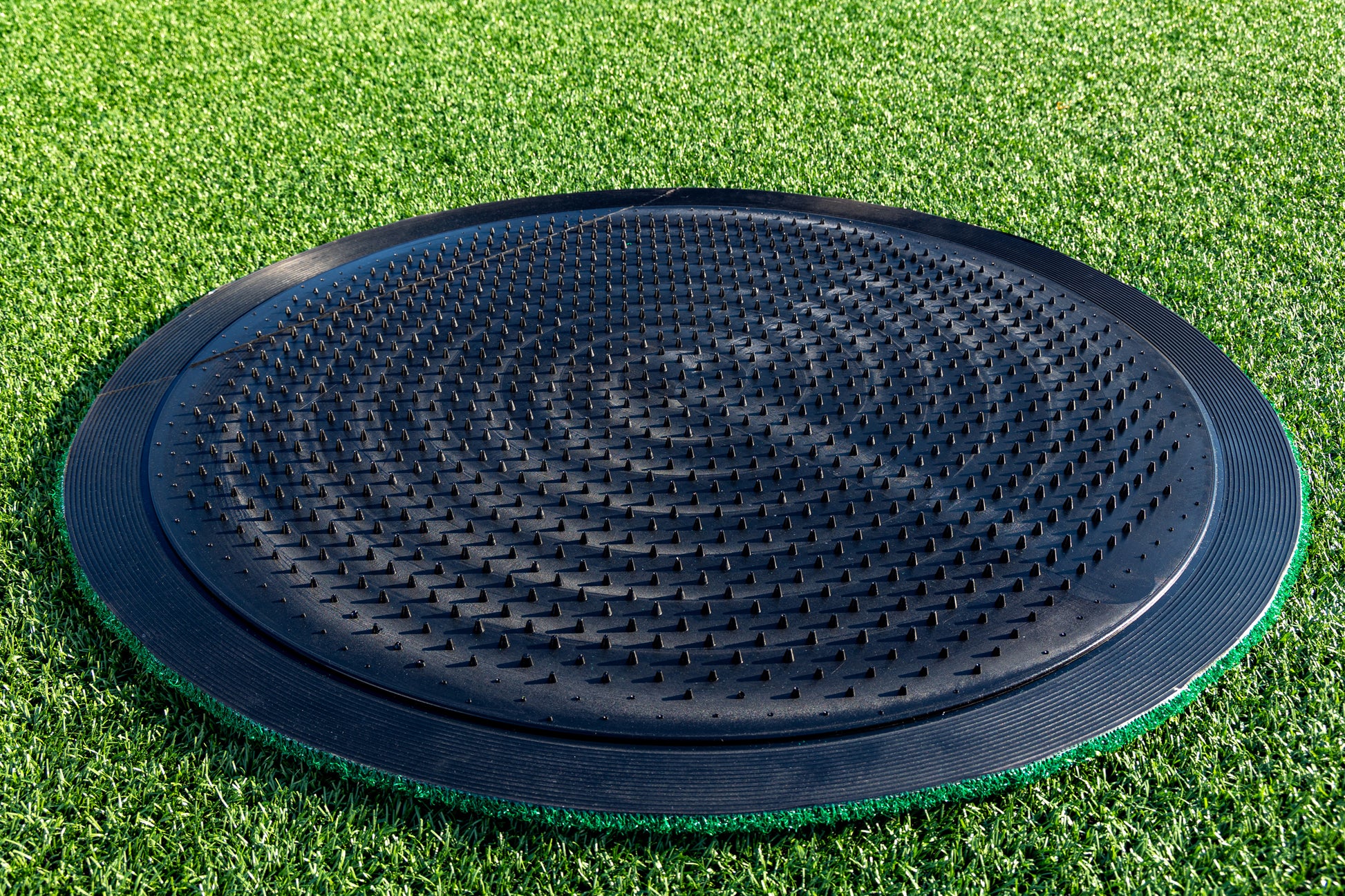 A Portolite On-Deck Circle featuring a large, black, textured plastic design with raised bumps is positioned on synthetic turf baseball grass, likely intended for balance or agility training near the on-deck area.