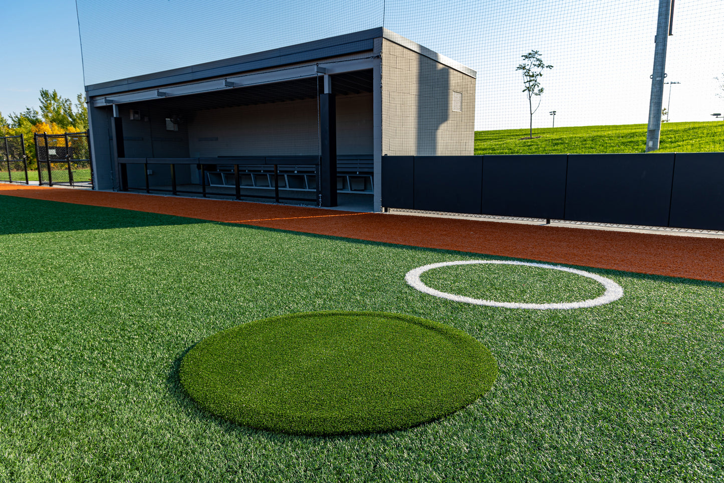 A baseball dugout with empty benches overlooks a vibrant synthetic turf baseball field. A pitcher’s mound and a Portolite On-Deck Circle—which is available in 4′, 5′, and 6′ sizes—are visible in the foreground, all brightly lit beneath clear skies.