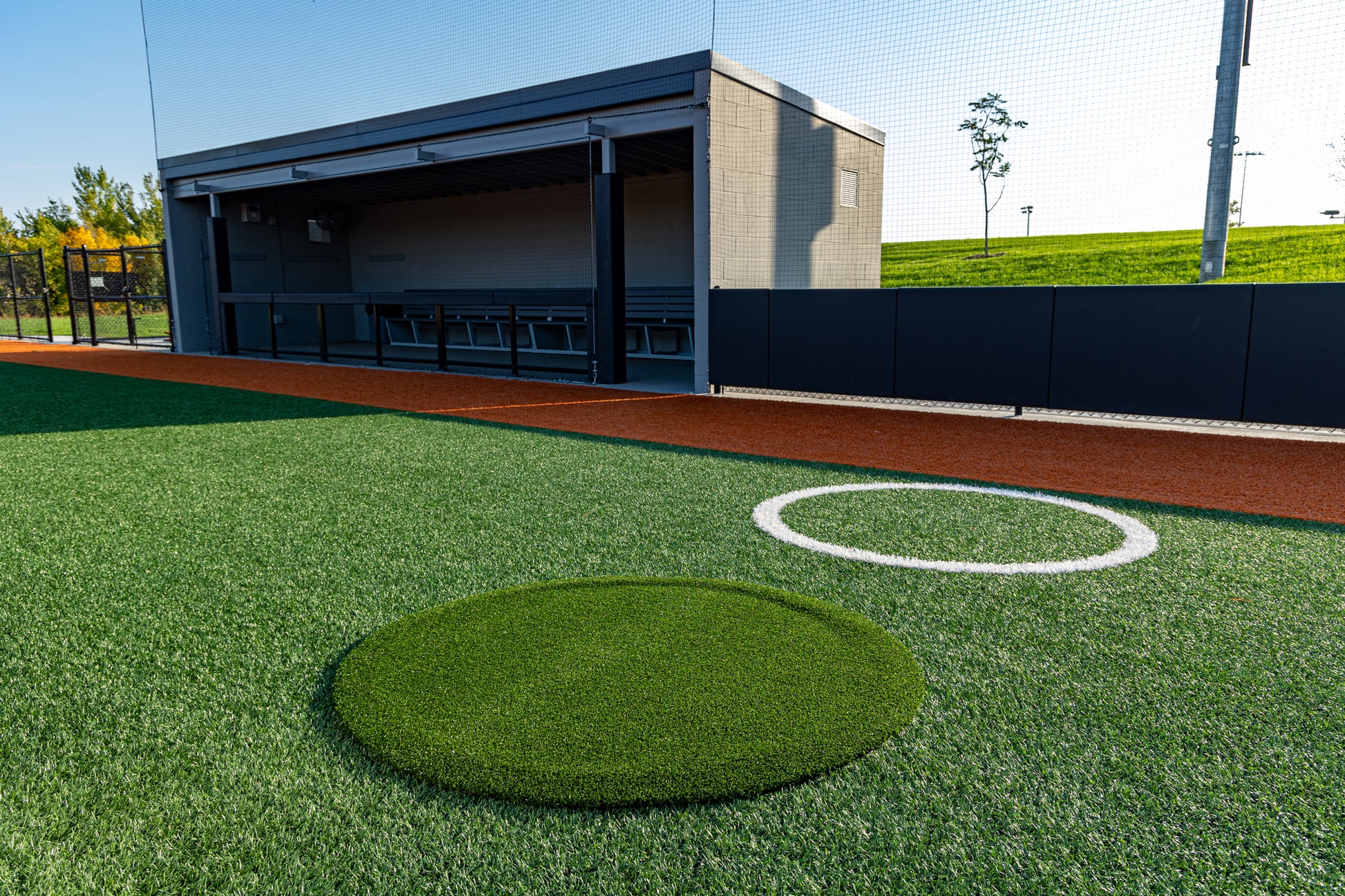 A baseball dugout with empty benches overlooks a vibrant synthetic turf baseball field. A pitcher’s mound and a Portolite On-Deck Circle—which is available in 4′, 5′, and 6′ sizes—are visible in the foreground, all brightly lit beneath clear skies.