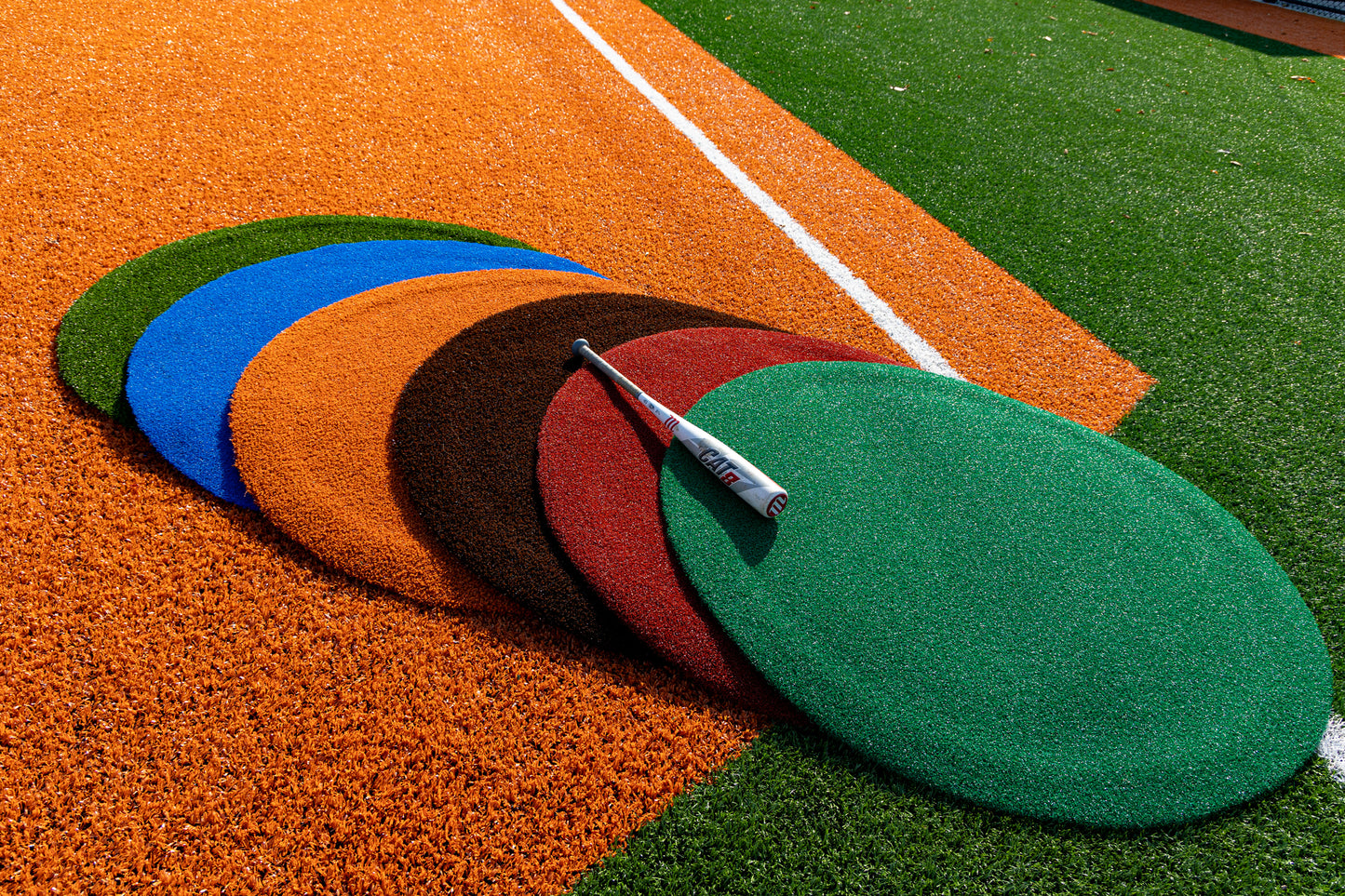 Five large, round Portolite On-Deck Circles in green, brown, red, orange, and blue lay overlapping on the synthetic turf baseball field. A baseball bat rests diagonally across the mats—suggesting an on-deck circle. White lines mark the field.