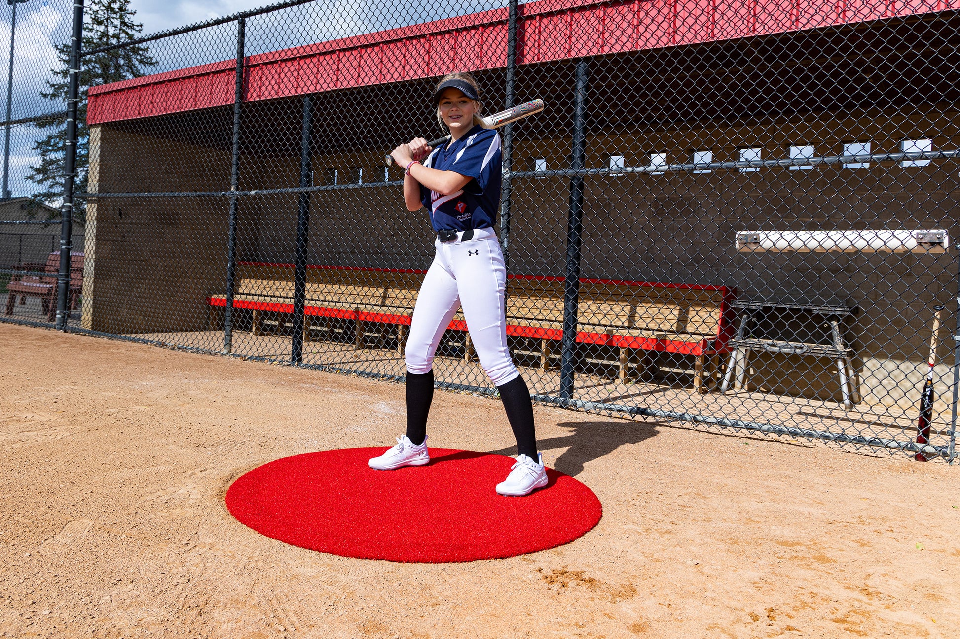 A baseball player in uniform stands on a Portolite On-Deck Circle, bat poised to swing, positioned in front of a fenced dugout on a sunny baseball field.