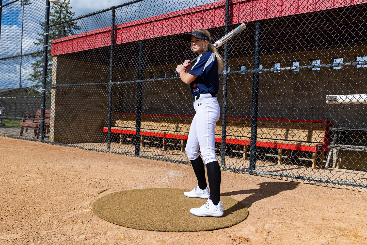 A softball player in uniform stands poised to bat near a dugout, holding a bat over her shoulder on a sunny day. Just beside the Portolite On-Deck Circle—available in 4′, 5′, and 6′ sizes—the dugout with its fenced front and red benches overlooks the well-maintained baseball field.