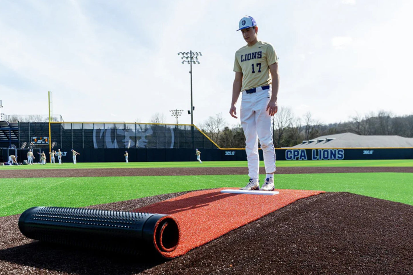 A baseball player in a Lions uniform stands on a white base on a turf field, with a Portolite Pitching Mound Protector | 13′ x 4′ Inlaid Pitching Mound Mat rolled up nearby and teammates practicing in the background.
