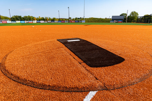 A bright orange baseball field featuring a pitchers mound equipped with the Portolite Pitching Mound Protector | 13′ x 4′ Inlaid Pitching Mound Mat and a home plate area, surrounded by green grass, trees, and advertisement signs along the outfield fence under a clear blue sky.