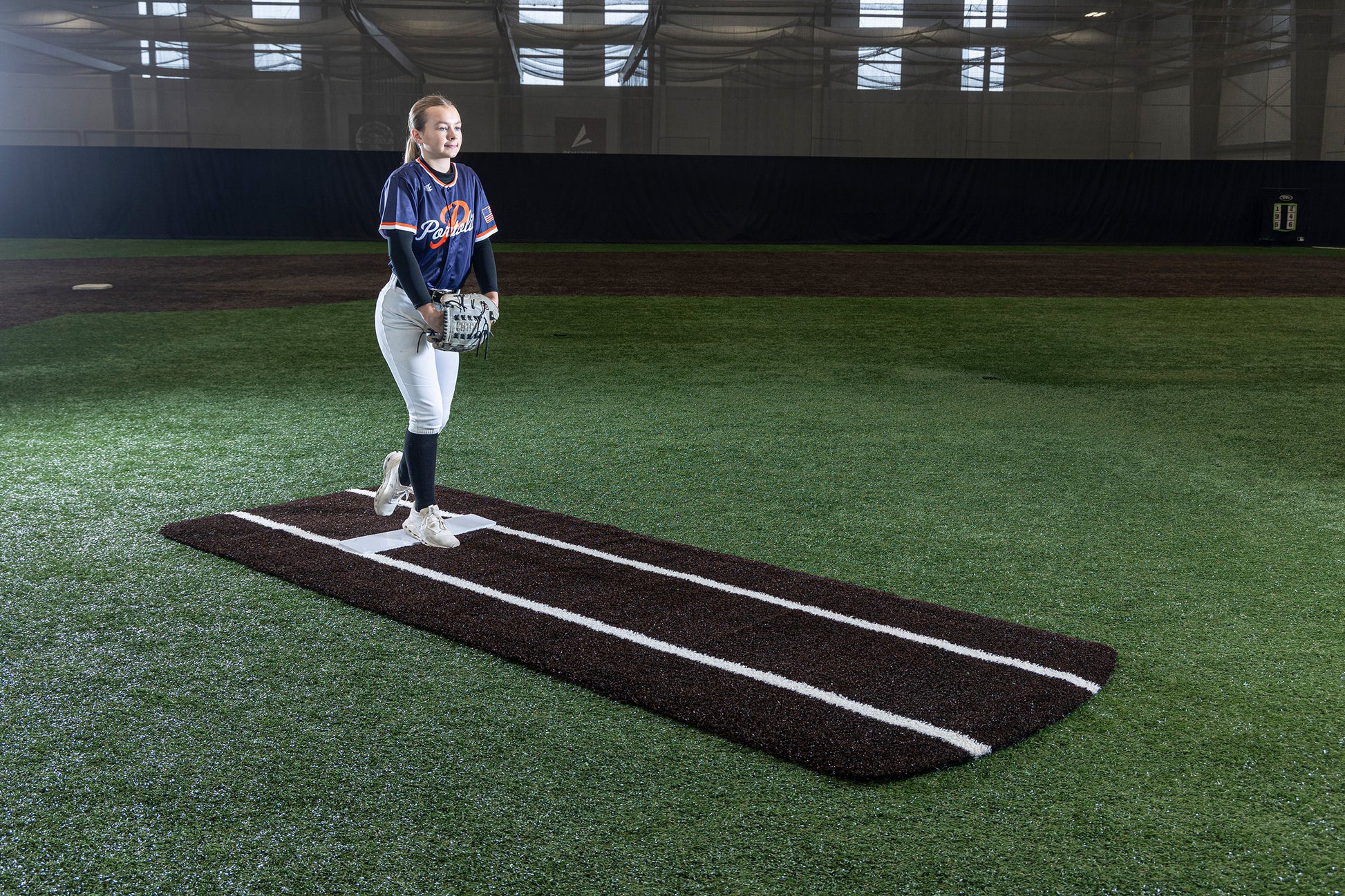 A softball pitcher stands on a Portolite Pro Spiked Game Mat with Lines / Lanes—built for collegiate and pro-level pitching—atop an indoor mound, wearing a dark jersey and white pants, glove in hand as she prepares to throw. The field beneath her is covered in green artificial turf.