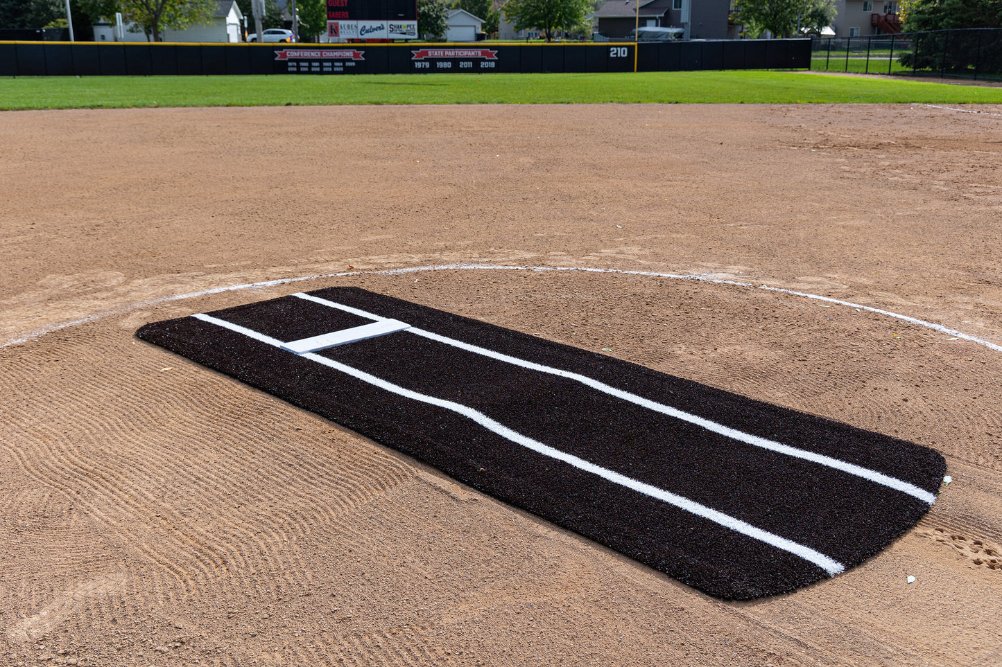 A Portolite Pro Spiked Game Mat with Lines / Lanes, built for collegiate and pro-level pitching, is placed on the pitcher’s mound of a baseball field. The black mat with white lines delivers excellent durability and improves field maintenance. In the background, you can see an outfield fence, scoreboard, and houses.