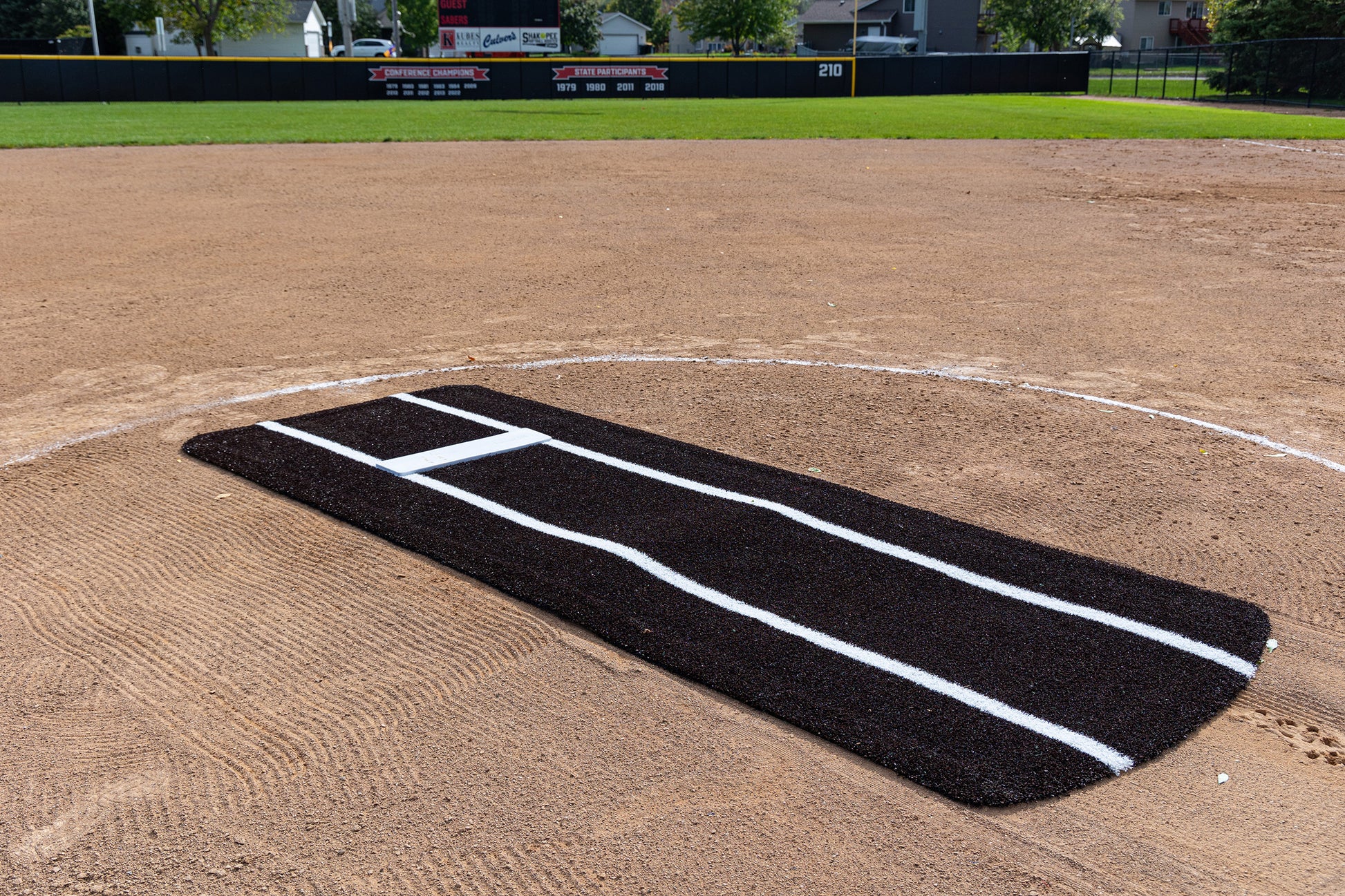 A Portolite Pro Spiked Game Mat with Lines / Lanes, built for collegiate and pro-level pitching, is placed on the pitcher’s mound of a baseball field. The black mat with white lines delivers excellent durability and improves field maintenance. In the background, you can see an outfield fence, scoreboard, and houses.