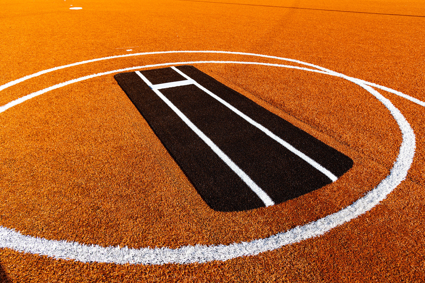 Orange cricket pitch with a black rectangular batting crease in the center, surrounded by a large white circle outlining the playing area. White lines mark the crease on the artificial turf, similar to those featured on the Portolite Pro Spiked Game Mat with Lines / Lanes, which is built for collegiate and pro-level pitching.