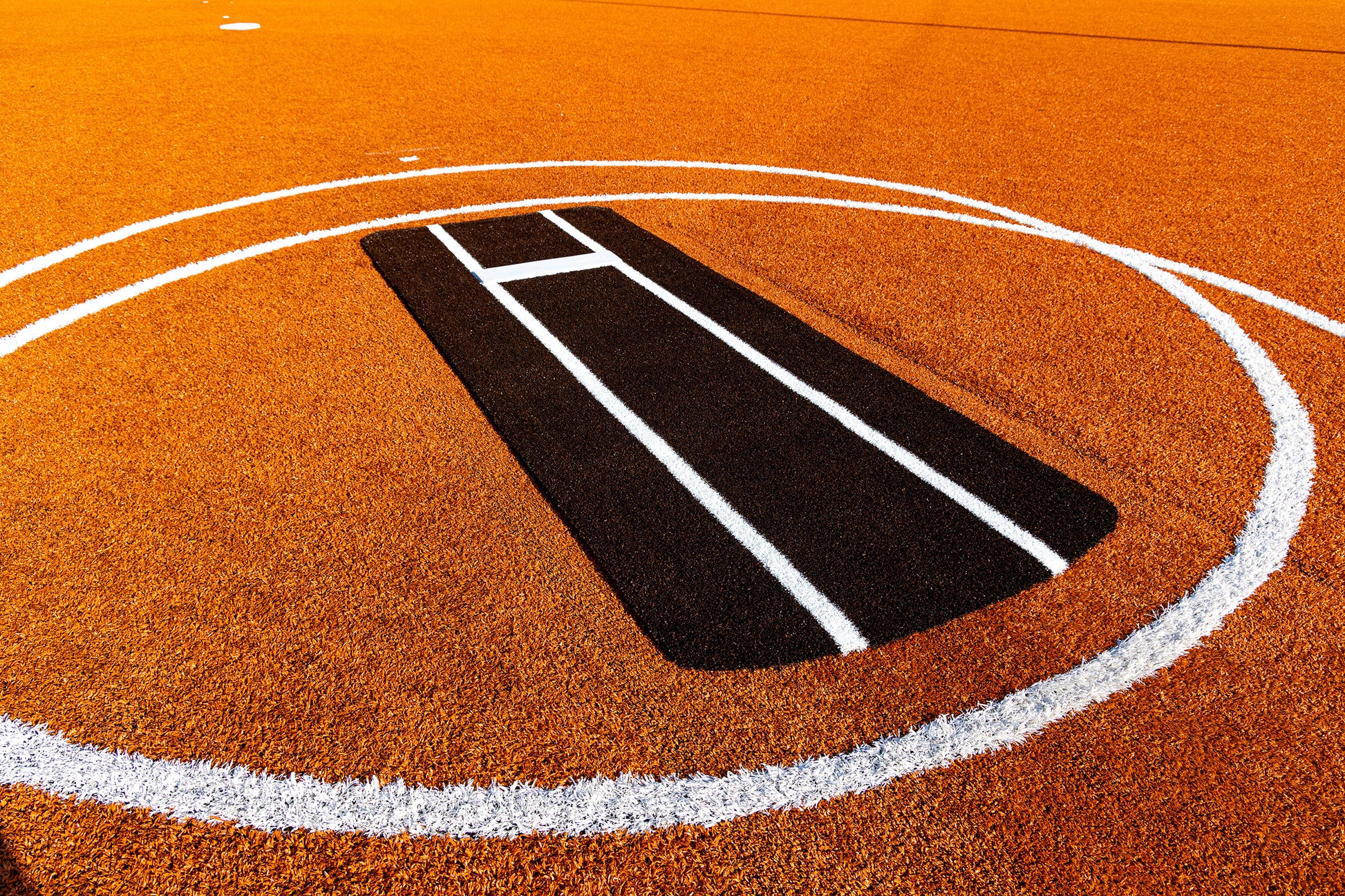 Orange cricket pitch with a black rectangular batting crease in the center, surrounded by a large white circle outlining the playing area. White lines mark the crease on the artificial turf, similar to those featured on the Portolite Pro Spiked Game Mat with Lines / Lanes, which is built for collegiate and pro-level pitching.
