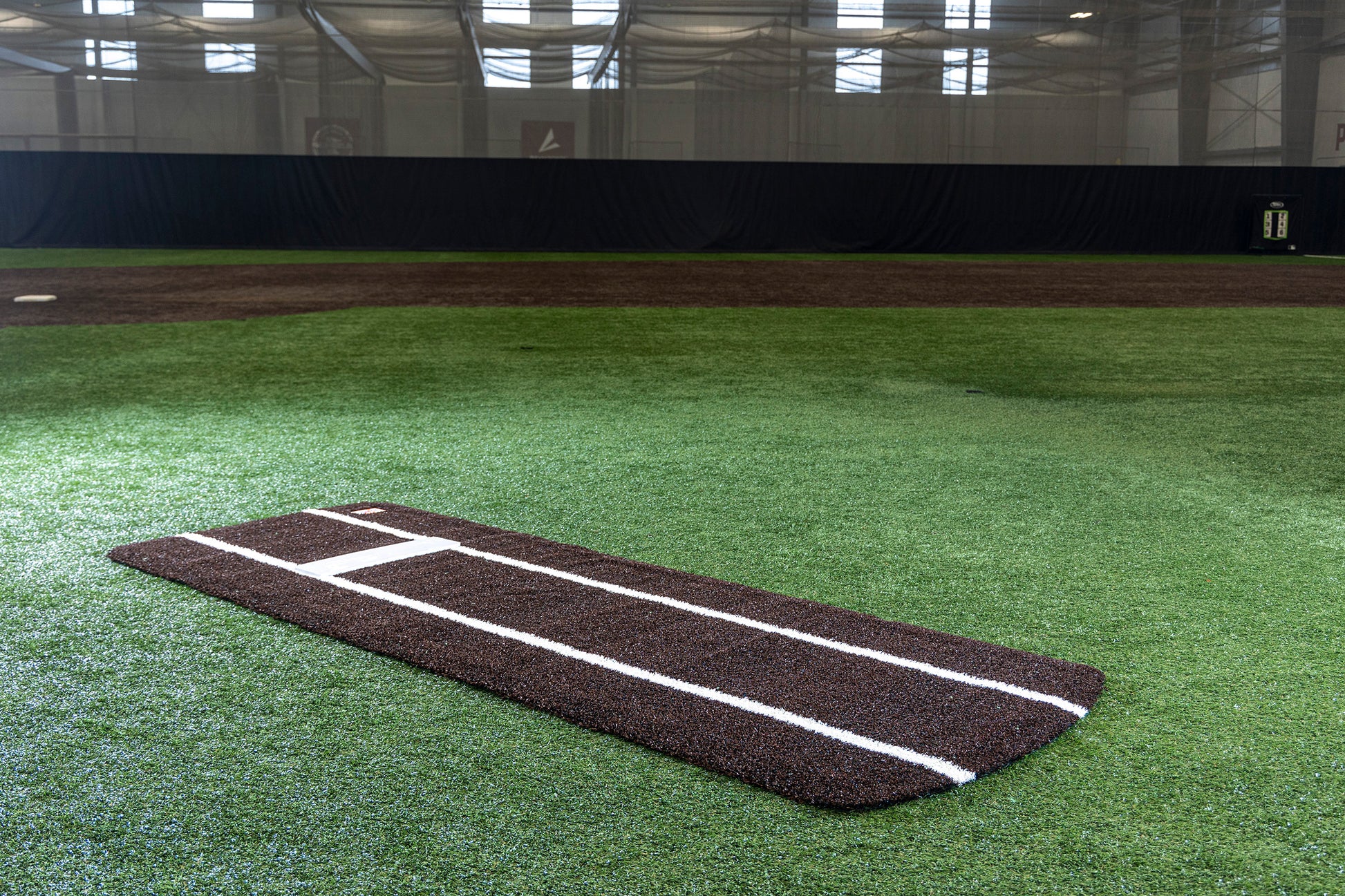 The Portolite Pro Spiked Game Mat with Lines / Lanes, designed for collegiate and pro-level pitching, in brown and white is placed on green artificial grass inside an indoor baseball training facility.