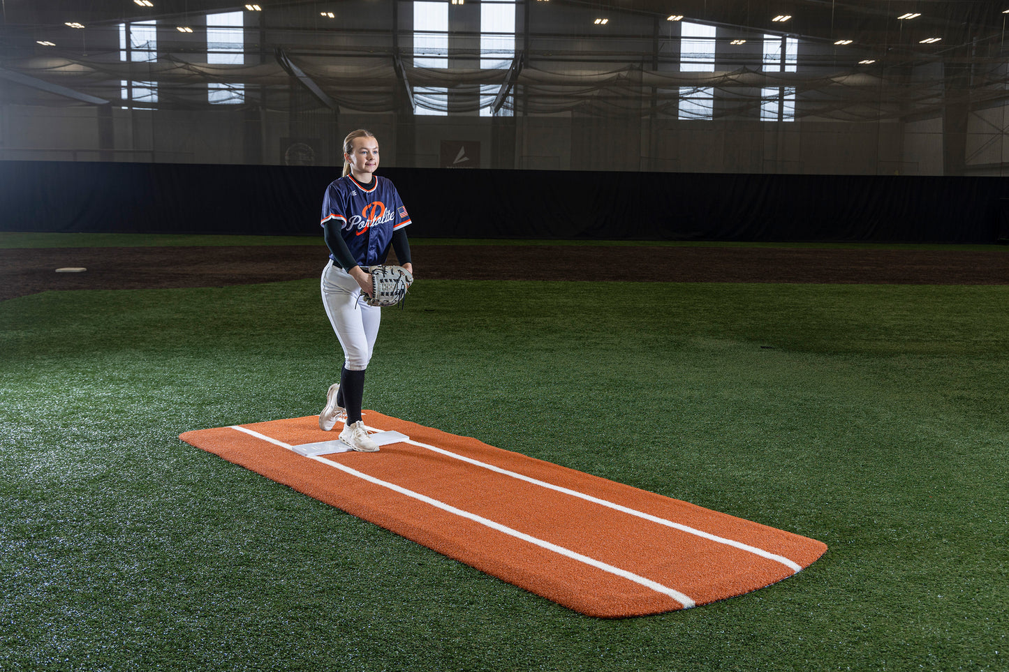 A softball player stands on the Portolite Pro Spiked Game Mat with Lines / Lanes—built for collegiate and pro-level pitching—inside an indoor field, wearing a navy blue jersey, white pants, and holding a glove. The well-maintained field is covered with artificial turf.