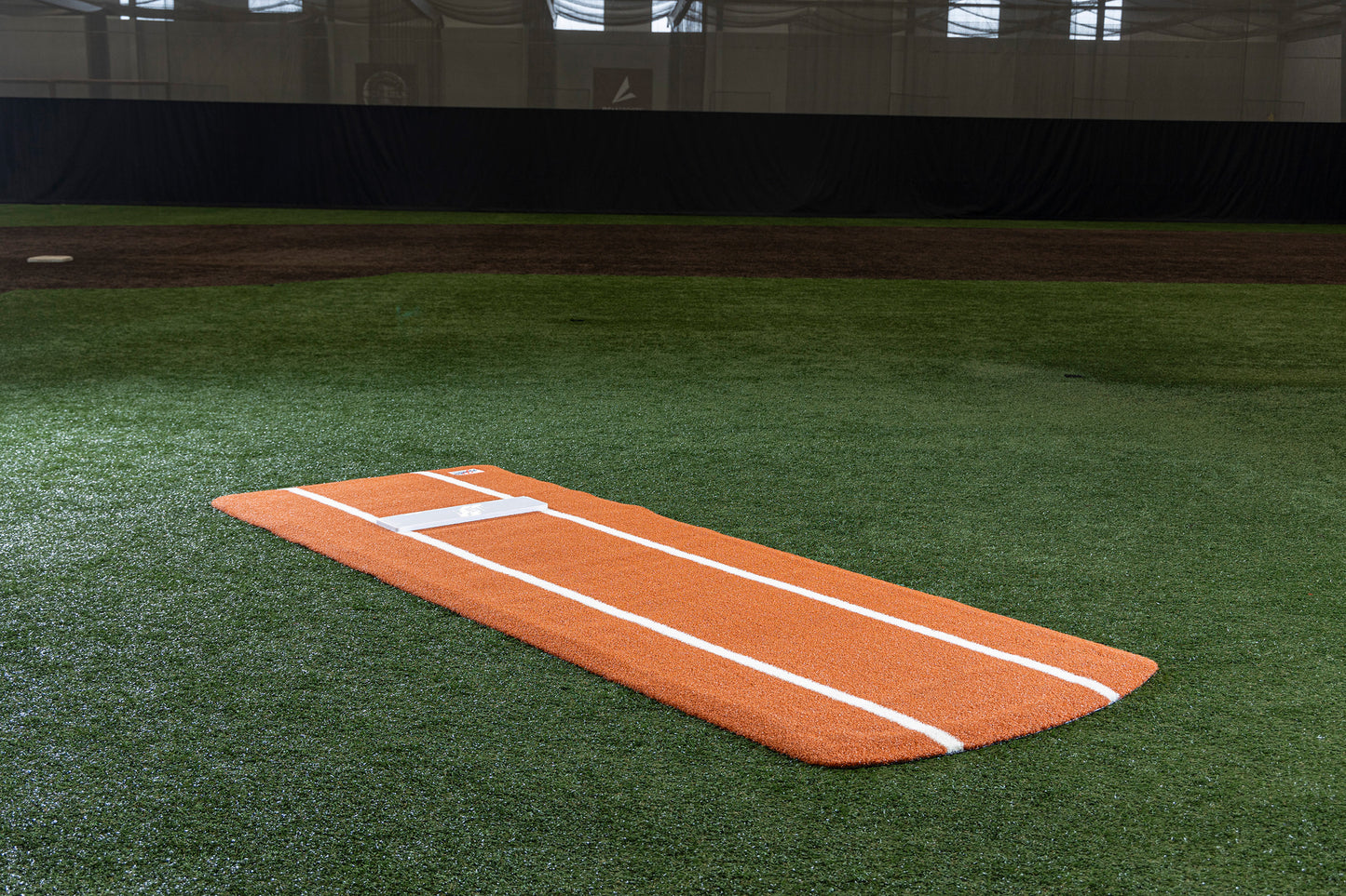 A Portolite Pro Spiked Game Mat with Lines/Lanes, built for collegiate and pro-level pitching, lies on green artificial turf inside an indoor baseball facility; its orange, portable mound with white lines provides excellent stability and makes field maintenance easy.