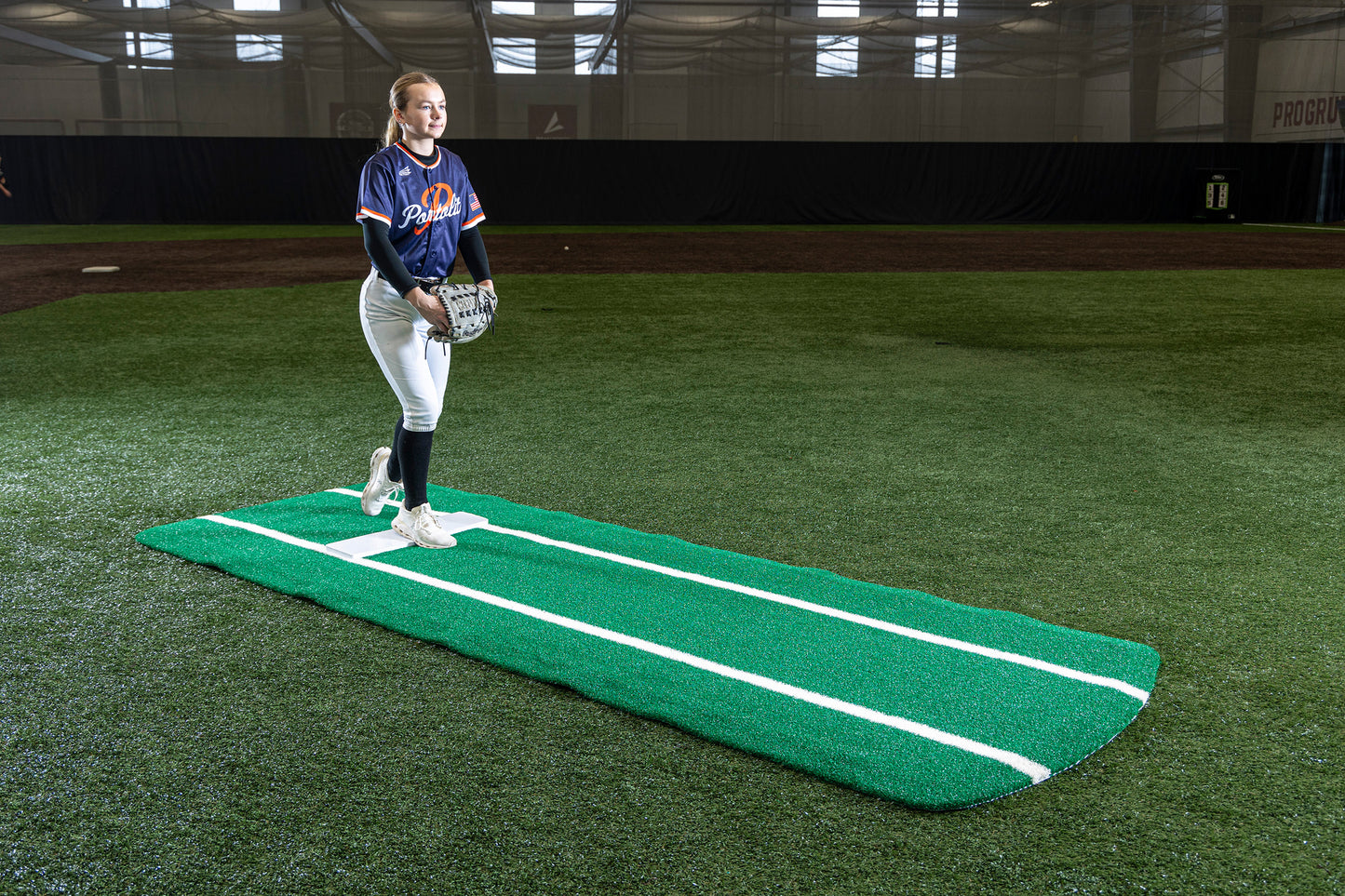 A softball player stands on a Portolite Pro Spiked Game Mat with Lines / Lanes—built for collegiate and pro-level pitching—inside an indoor field, wearing a blue jersey and white pants, holding a glove, and preparing to pitch.