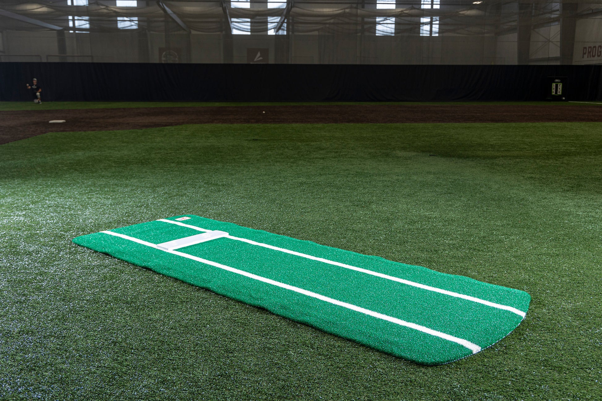 The Portolite Pro Spiked Game Mat with Lines / Lanes, built for collegiate and pro-level pitching, is shown in green with white lines on an indoor baseball field featuring turf, netting, and a dark backdrop. Designed for durability and optimal field maintenance during practice sessions.