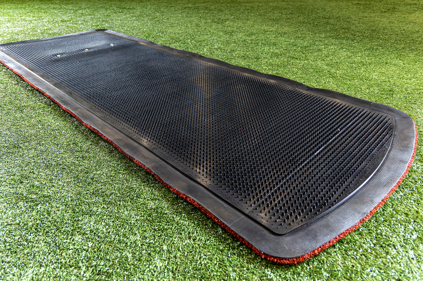 The Portolite Pro Spiked Game Mat with Lines / Lanes, designed for collegiate and pro-level pitching, features a large black rectangular shape with a textured surface, lying on green artificial turf and bordered by a thin strip of red material—ideal for field maintenance or as a top-quality softball pitching mat.